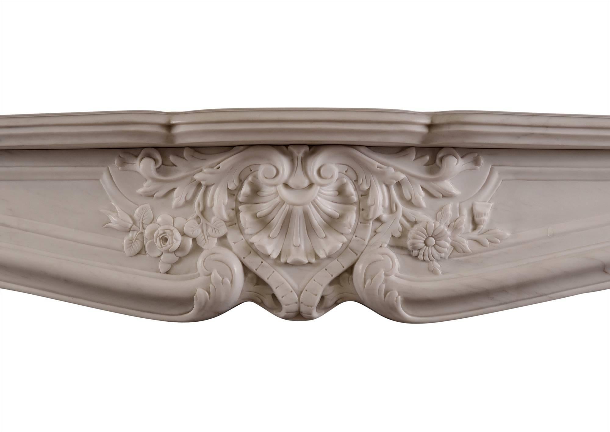 A richly carved Louis XV style fireplace in white marble. The flowing frieze with carved shell, flowers and foliage, shaped jambs with scroll and shell to top, leading the floral drops and stiff acanthus leaves below. Serpentine shaped shelf. The