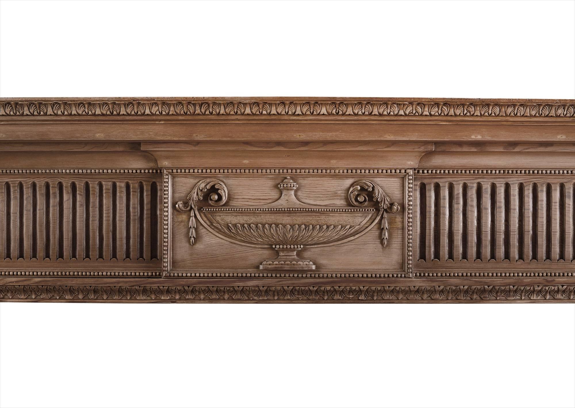 An English wood fireplace in the Georgian manner. The fluted frieze with carved urn to centre tablet adorned with carved beading, the tapering, panelled jambs with bellflowers and tied ribbons, surmounted Athenian leaf to end block. Moulded shelf