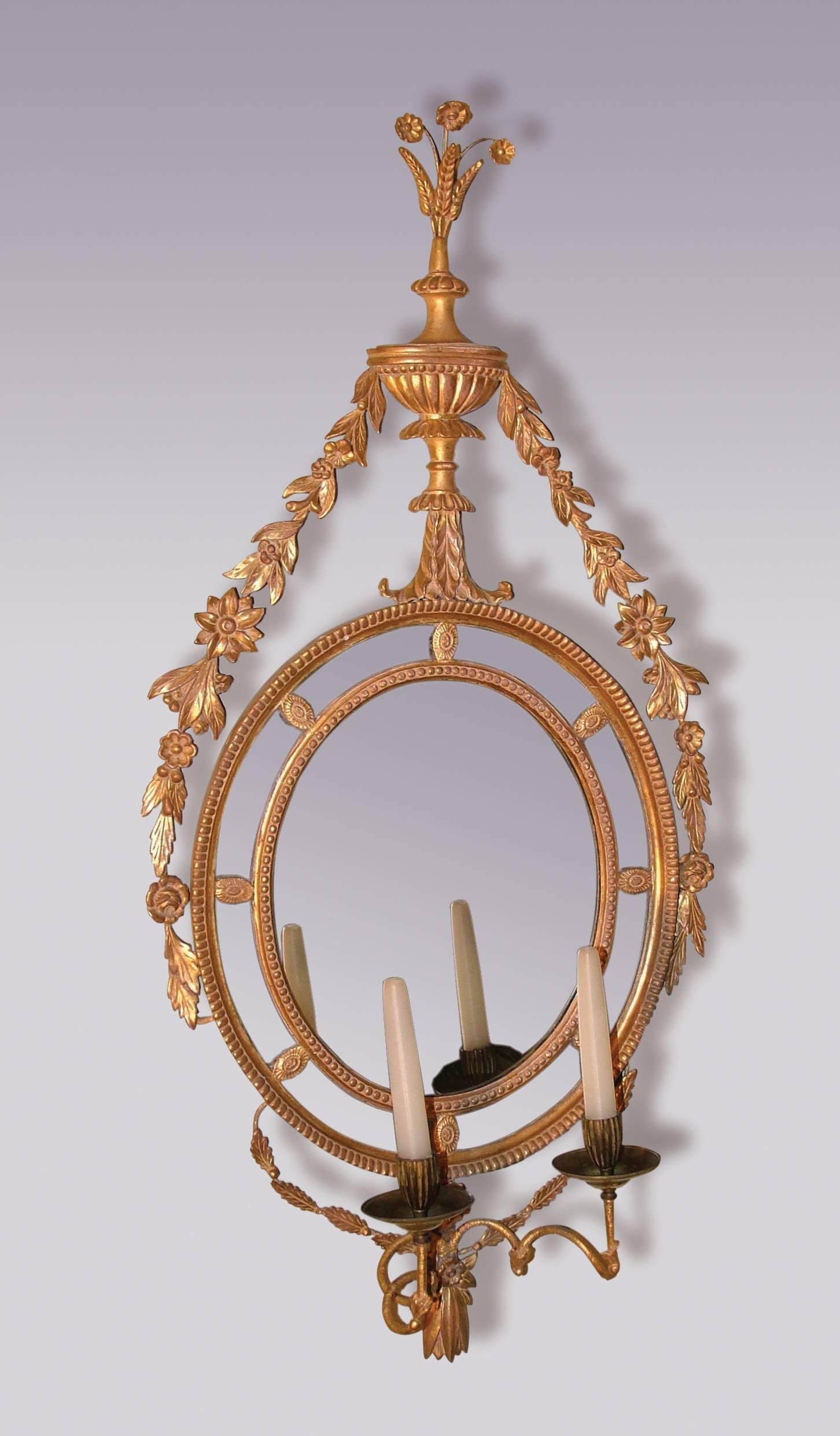 A pair of late 18th century Adam period giltwood two-light girandoles in the form of oval border glass mirrors with ornate leaf and flower carved swags, having urn pediments surmounted by wheat ear decoration. Some carvings and crestings possibly