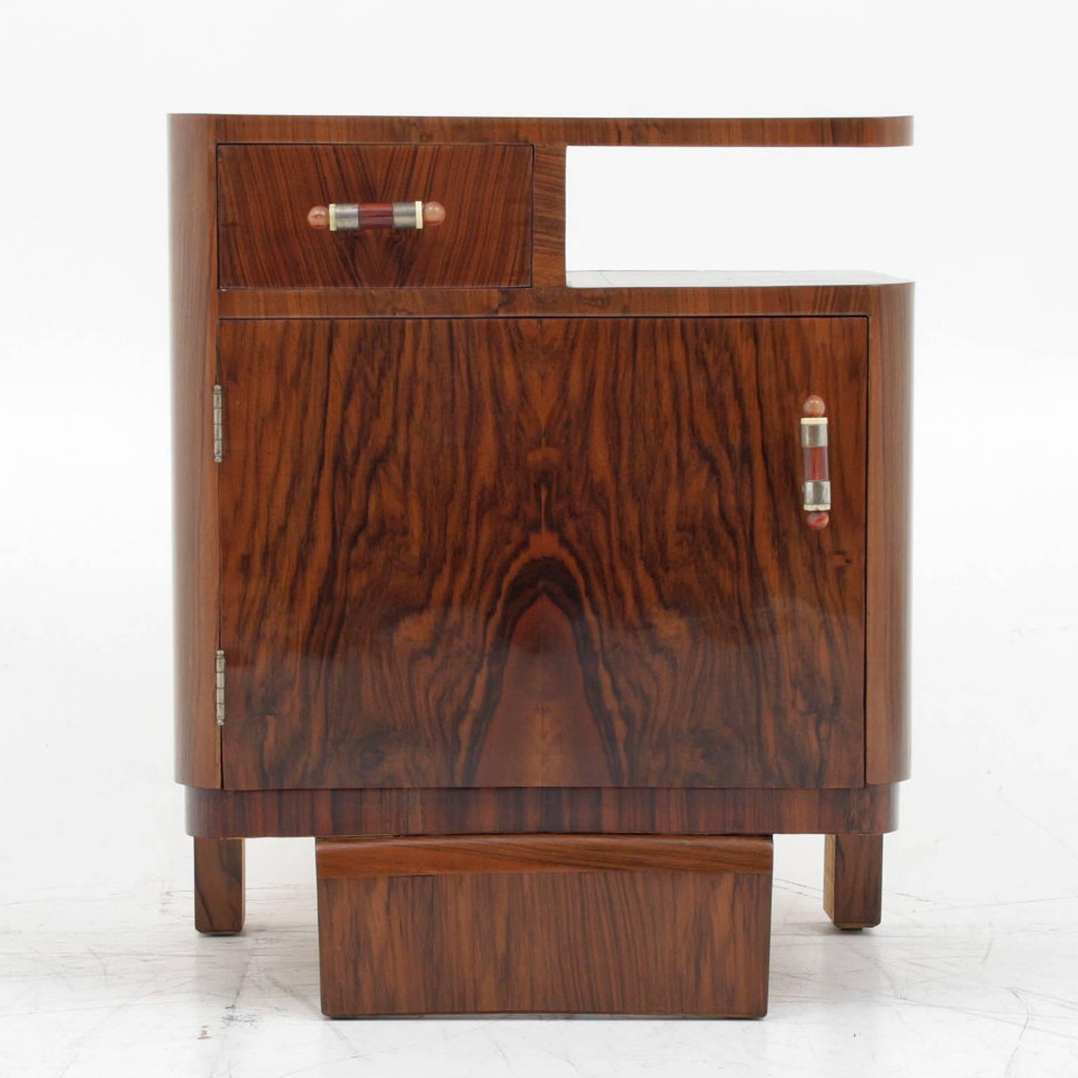 Two Art Deco bedside tables which have been formed to mirror each other. They stand on three legs, with the front one being wider than the other two. The tables have one small drawer at the top and in the middle of the body they have a larger