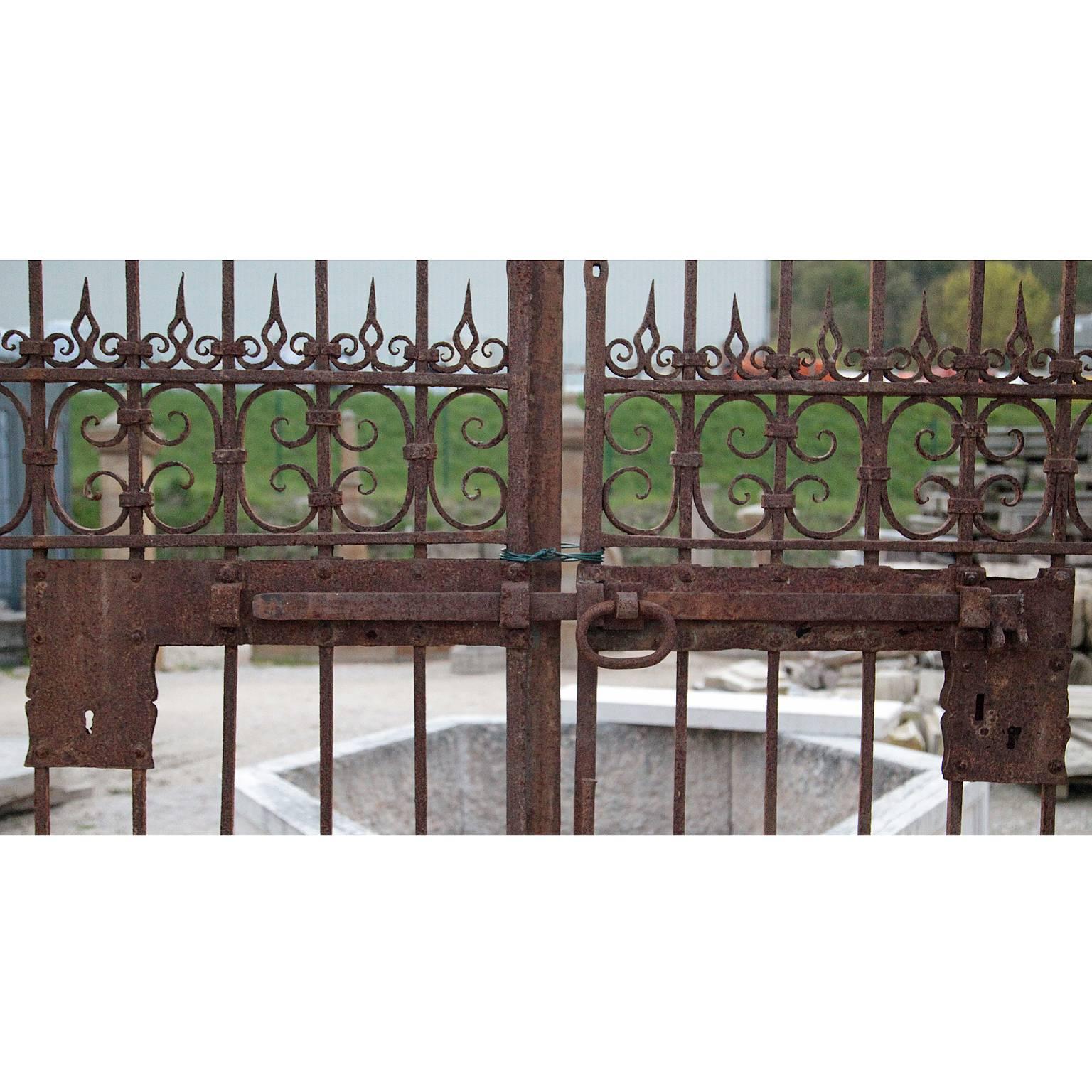 A large iron gate with C- and S-braces as well as flame-shaped elements which sit on top. The gate features two entry ways for people on either side and a large central, two-doored gate for car access.