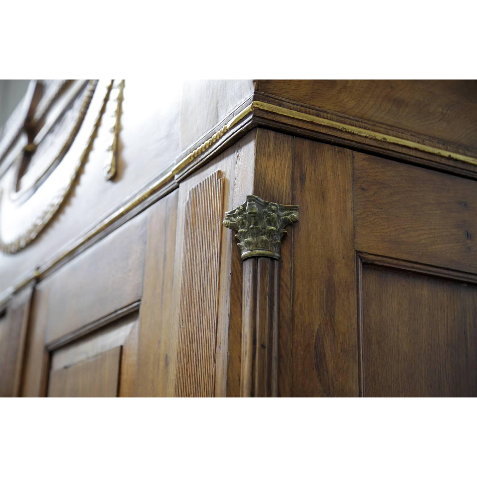 Large two-doored oak cupboard with Corinthian columns on the corners and dentils on doors, pediment and skirt. The pediment shows carved ornaments and two urns. The gilding was restored.