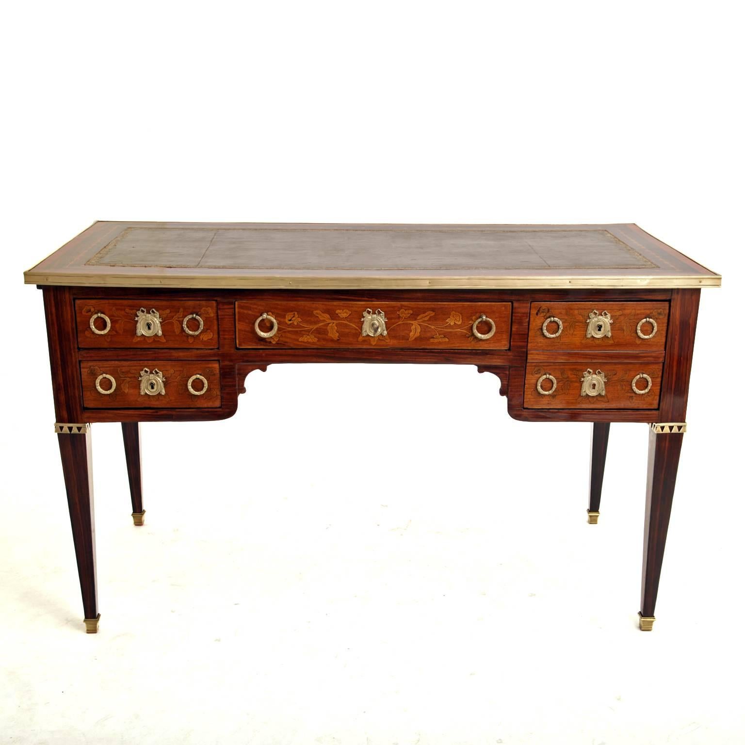 Bureau Plat in Louis-Seize-style on tapered feet with brass caps and five drawers. The edge of the tabletop is decorated with brass trim; the writing surface is covered in brown leather and inlaid with mahogany. The drawers show inlay work in the