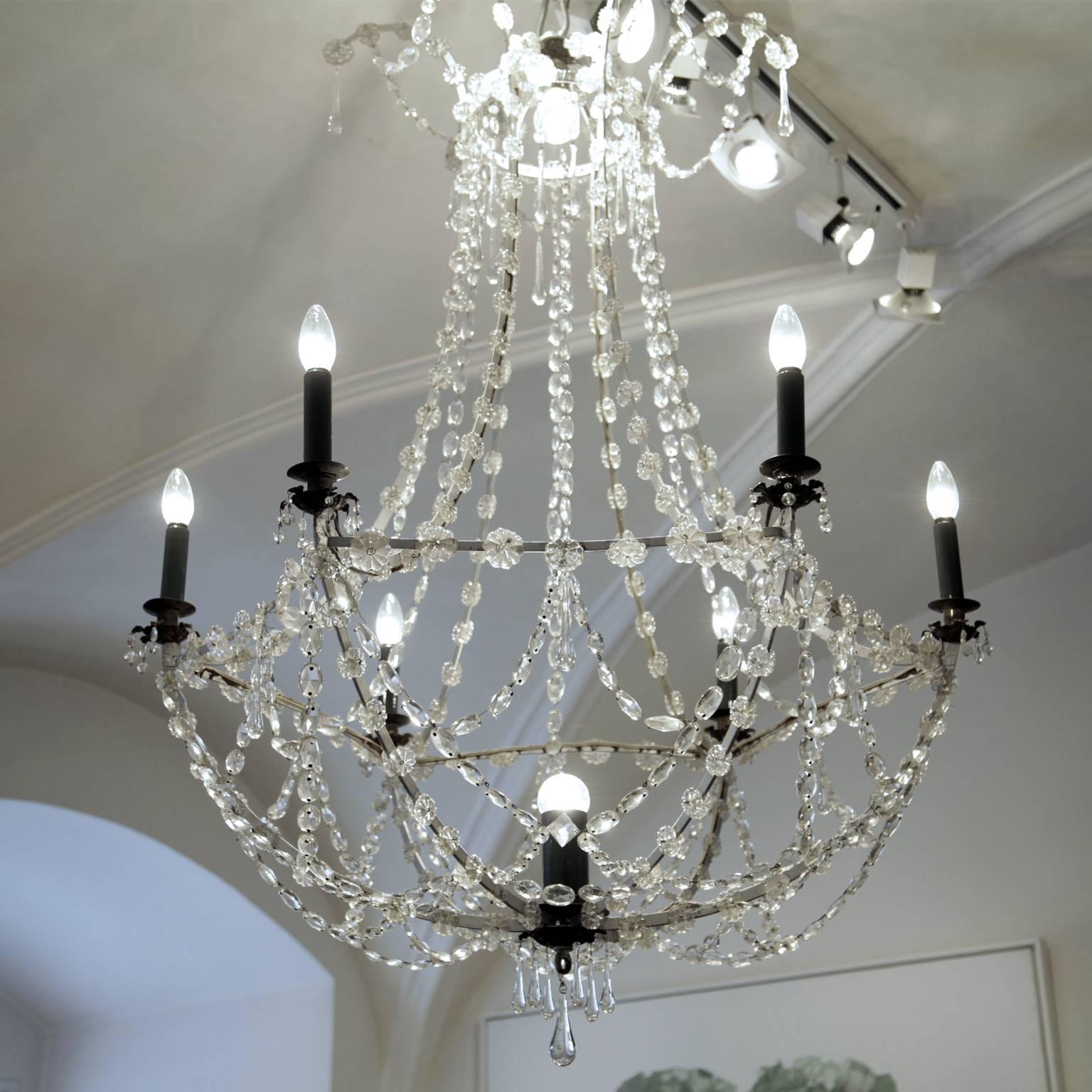 Glass chandelier with six lights around the gallery and one in the basket. The crown is decorated with hanging droplets while the basket ends in a droplet-shaped final. The candle slips are all black, which provides a nice contrast to the glass