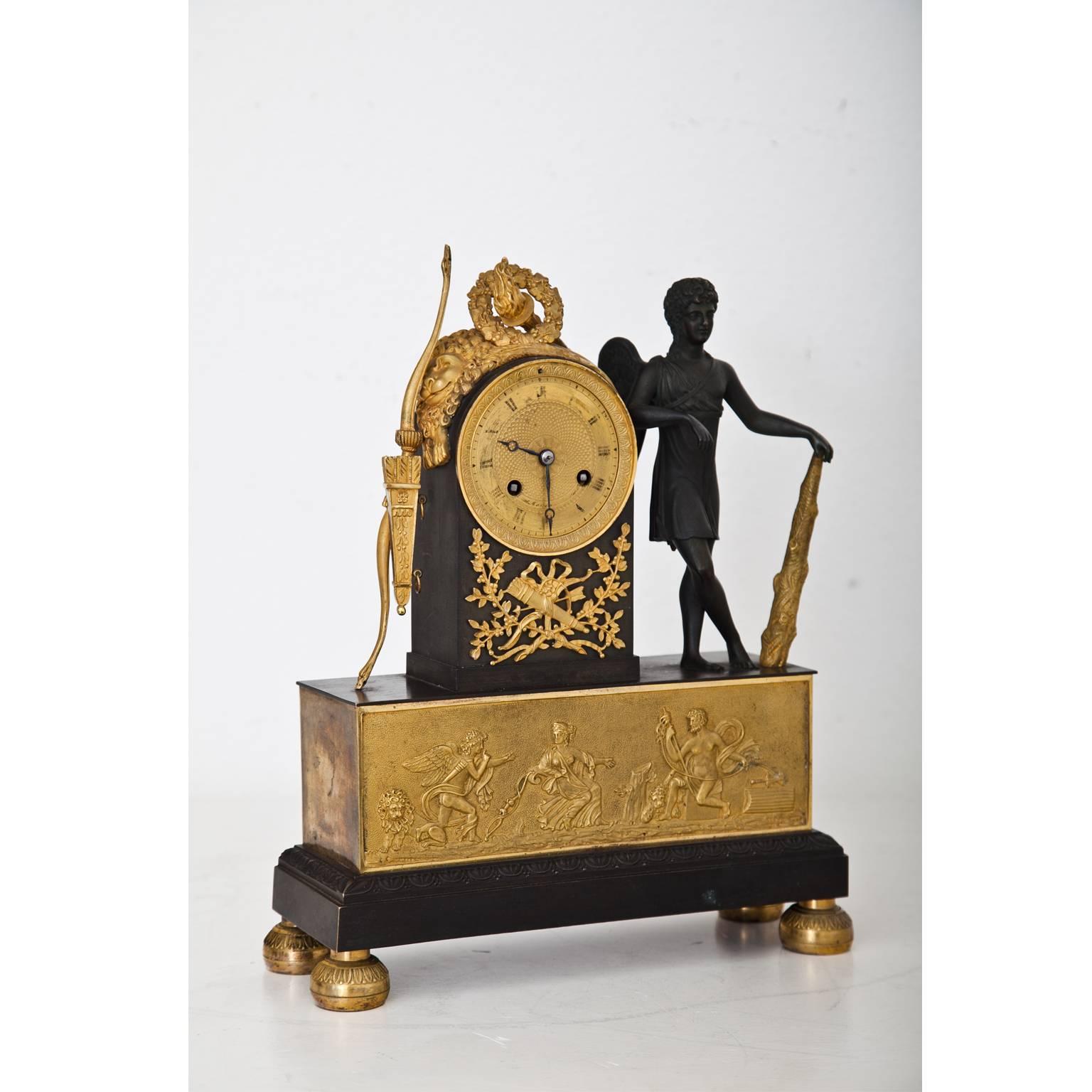 Pendule clock on low round feet with a rectangular base. The clockwork is in a rounded case, decorated with a torch, Hercules' lion skin and a laurel wreath. Next to it is the three-dimensional figure of cupid with Hercules' club and on the left