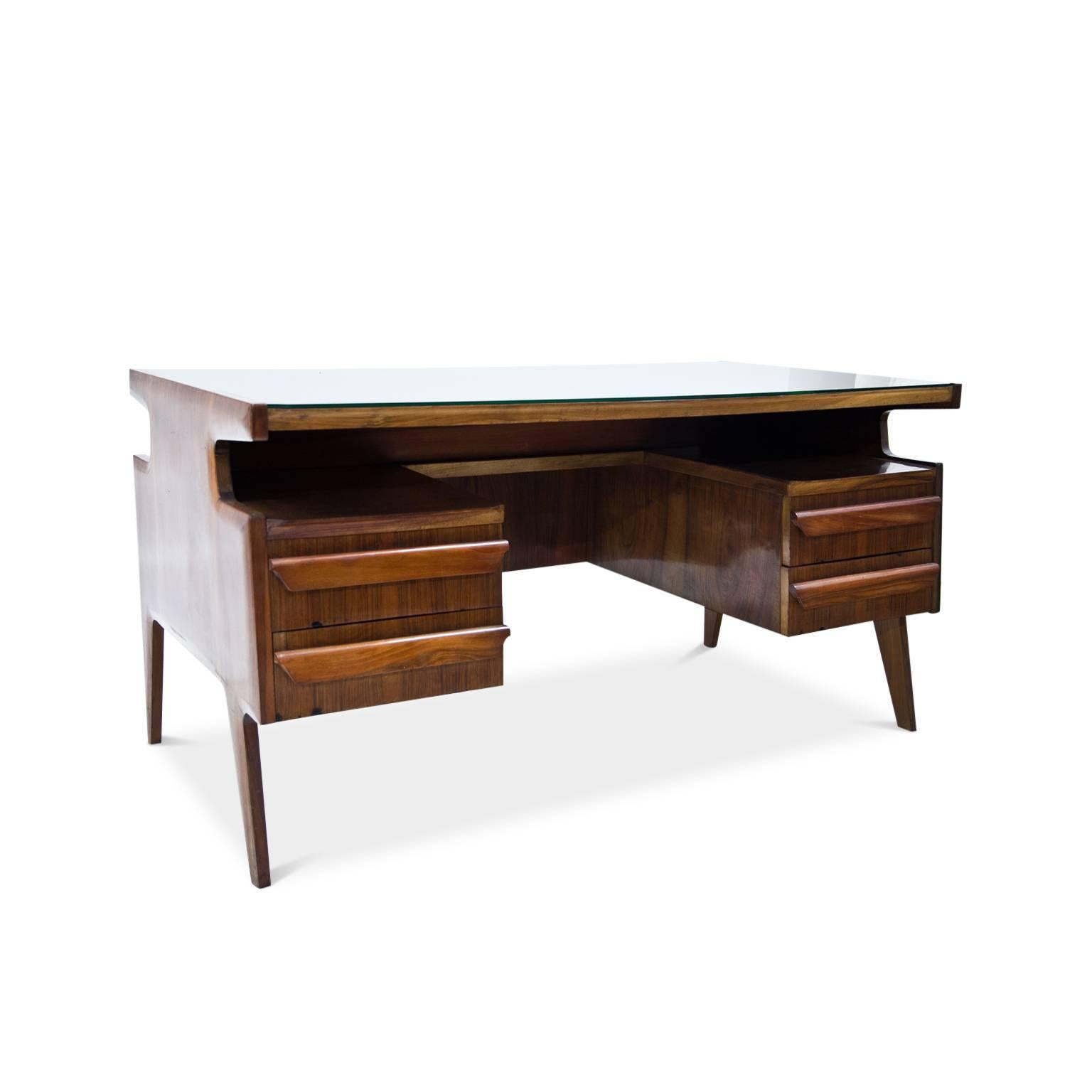 Elegant executive desk on slightly angled legs. The writing surface is covered with a green-tinted glass pane. Underneath is a shelving area and four drawers. The table shows a beautiful veneer pattern and is designed for an all-round view.