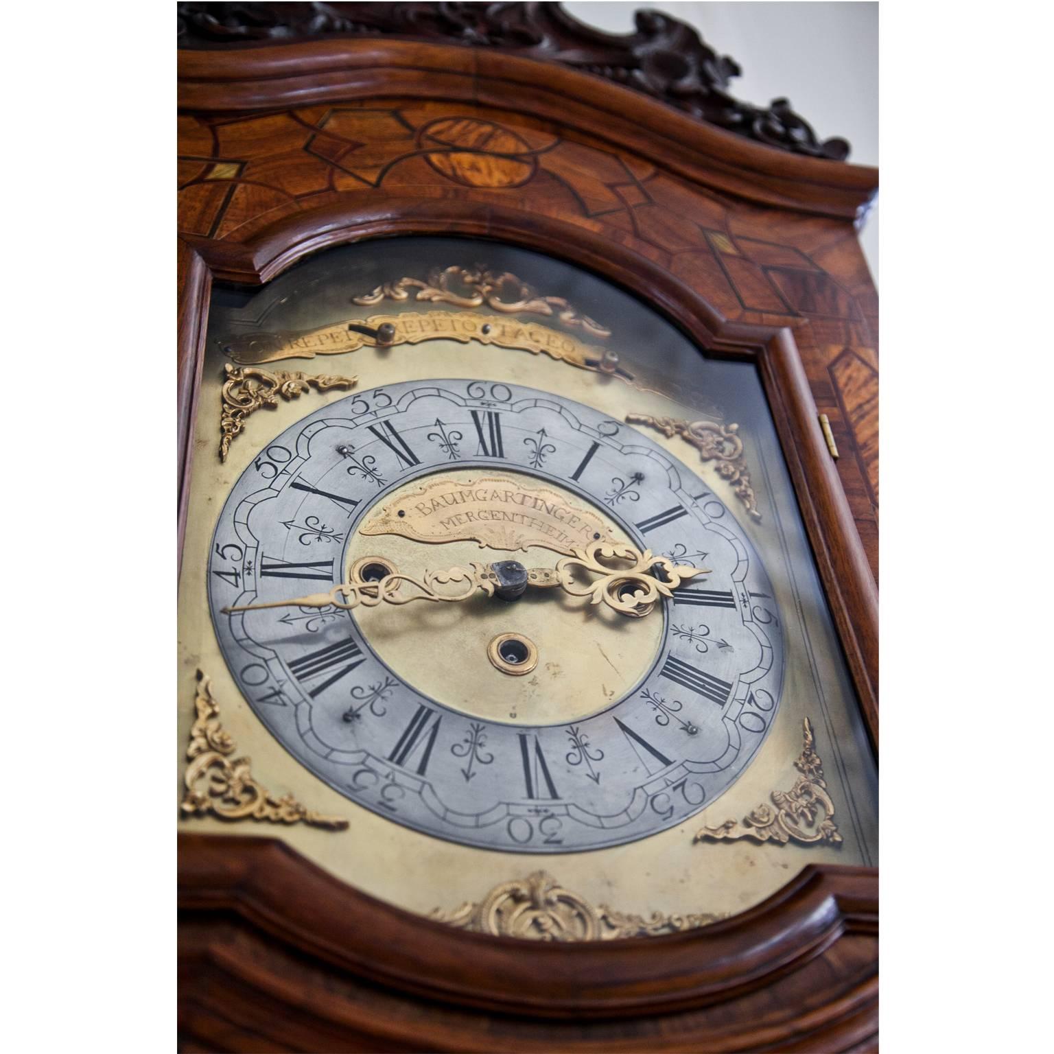 Grandfather clock with carved top and decorated with inlaywork. The clockface with Roman numerals reads Non Repet Repeto Taceo Audio and Baumgartinger Mergentheim.