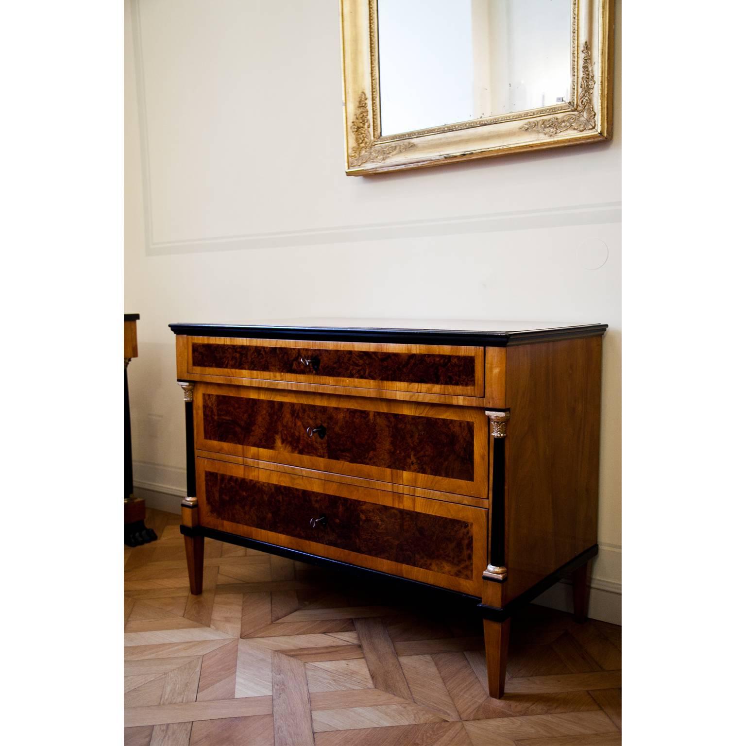 Three-drawered Biedermeier chest of drawers on tapered legs. The streamlined body shows ebonized and partly gilt columns, supporting the slightly protruding top drawer. The front is inlaid with burl wood veneer and the edges are ebonized as well.