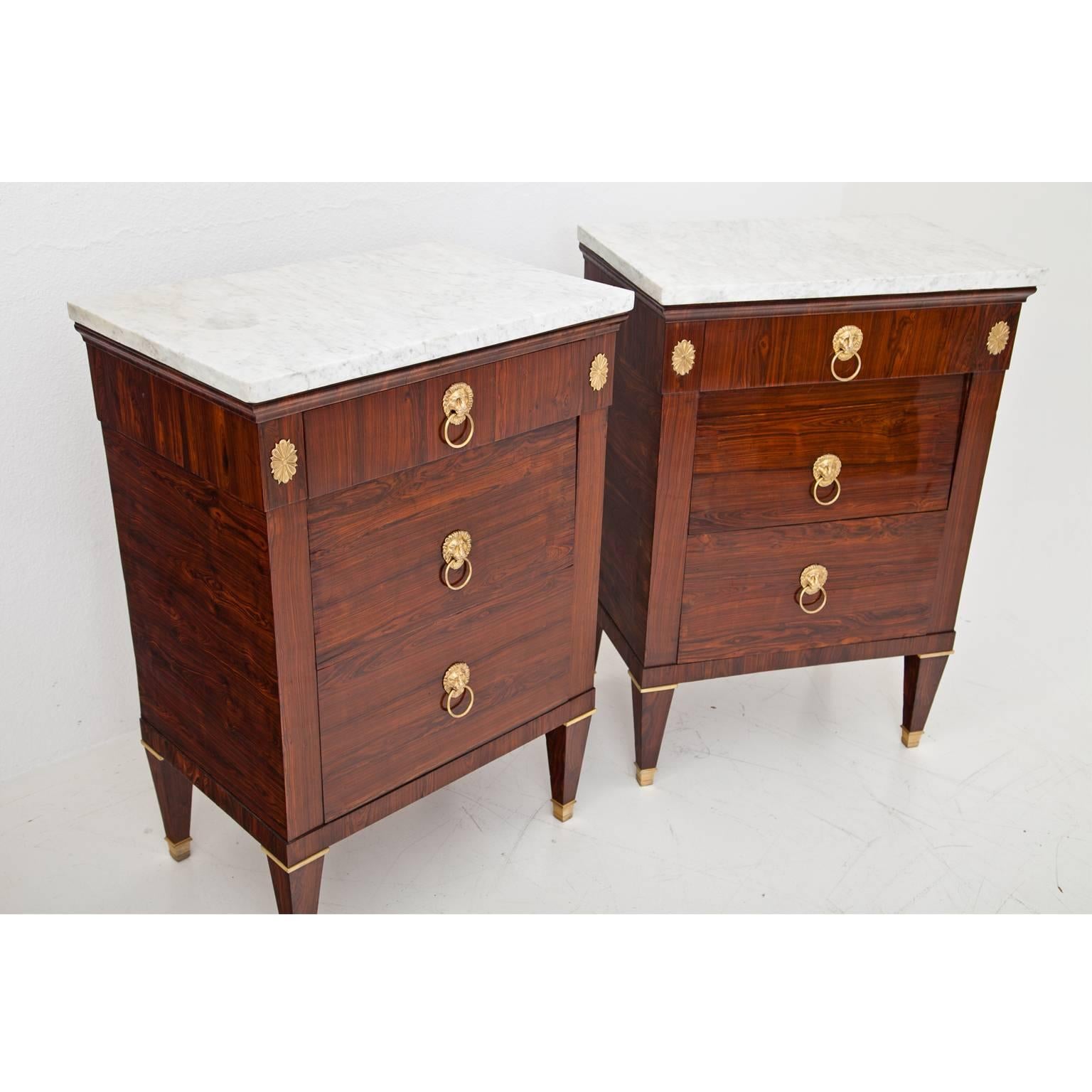 Pair of neoclassical chests of drawers on tapered feet with brass sabots, a white marble top and lionhead fittings. The chests each have a top drawer and a middle compartment with a folding door. The lowest drawer is designed as a pull-out trunk