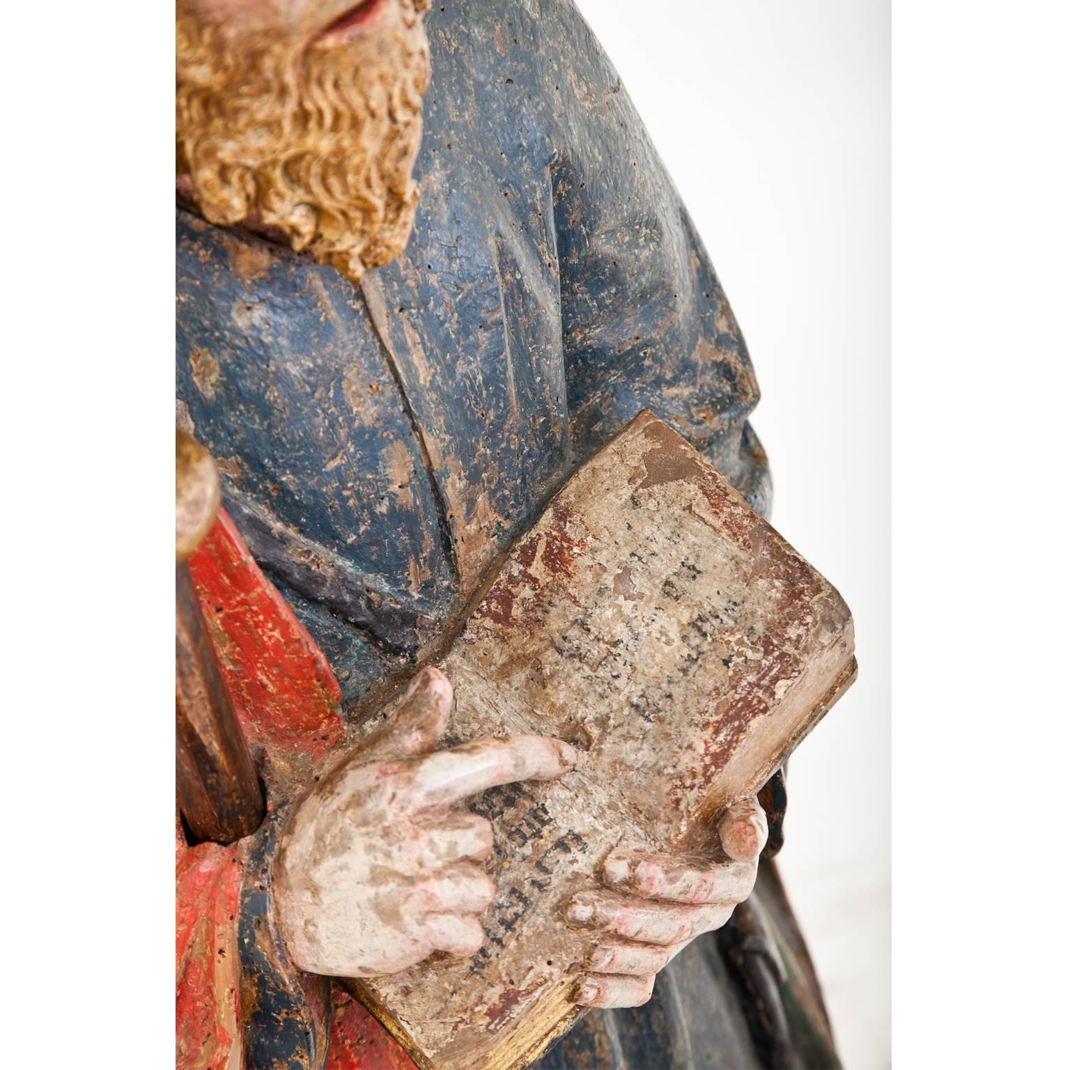 Large sculpture of Saint James with his walking staff and his attribute of the scallop on his hat. He holds in his hands a book and with his right index fingers points to a text passage. He is depicted as barefoot, wearing a blue garment and a red