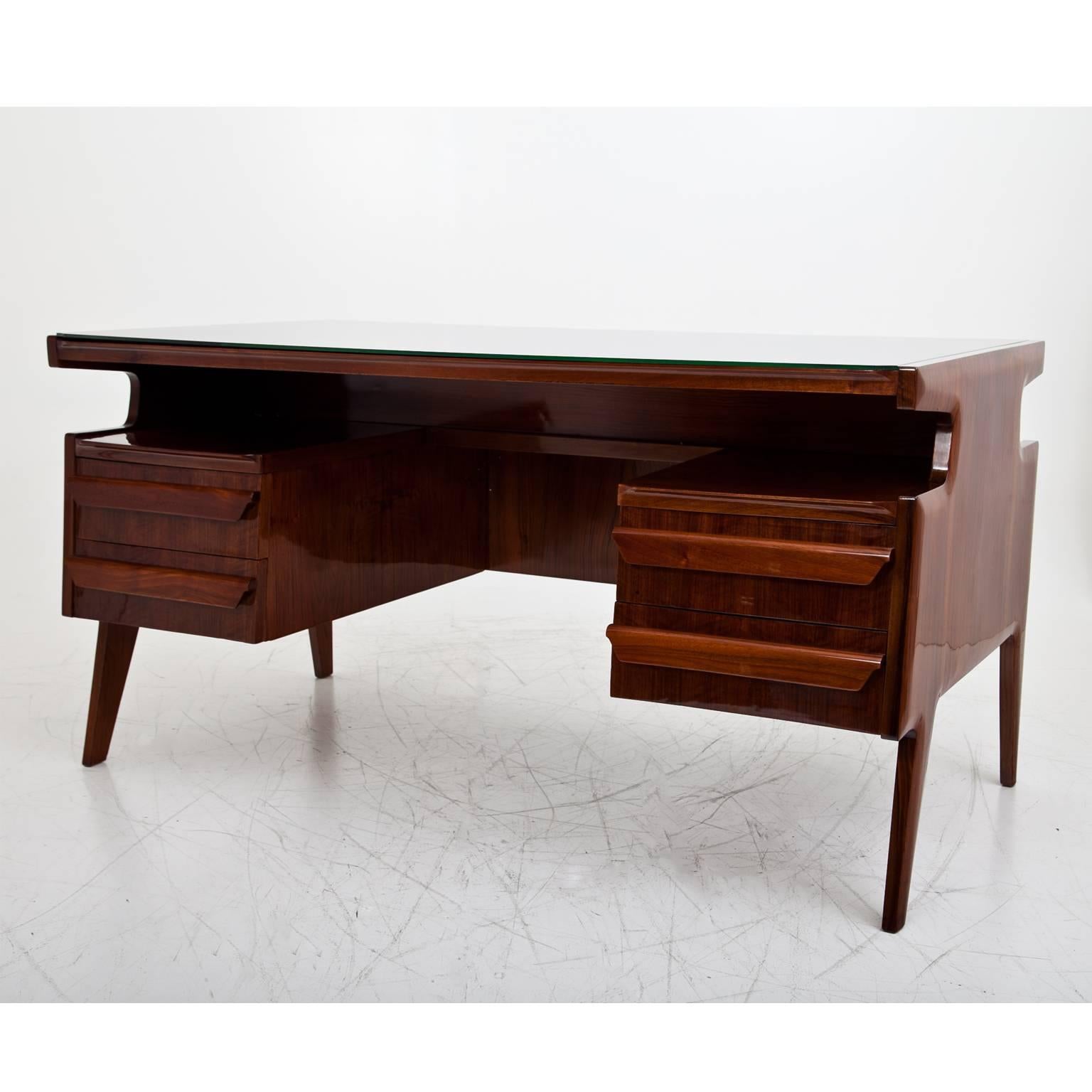 Elegant executive desk on slightly angled legs. The writing surface is covered with a green-tinted glass pane. Underneath is a shelving area and four drawers. The table shows a beautiful veneer pattern and is designed for an all-round view. The