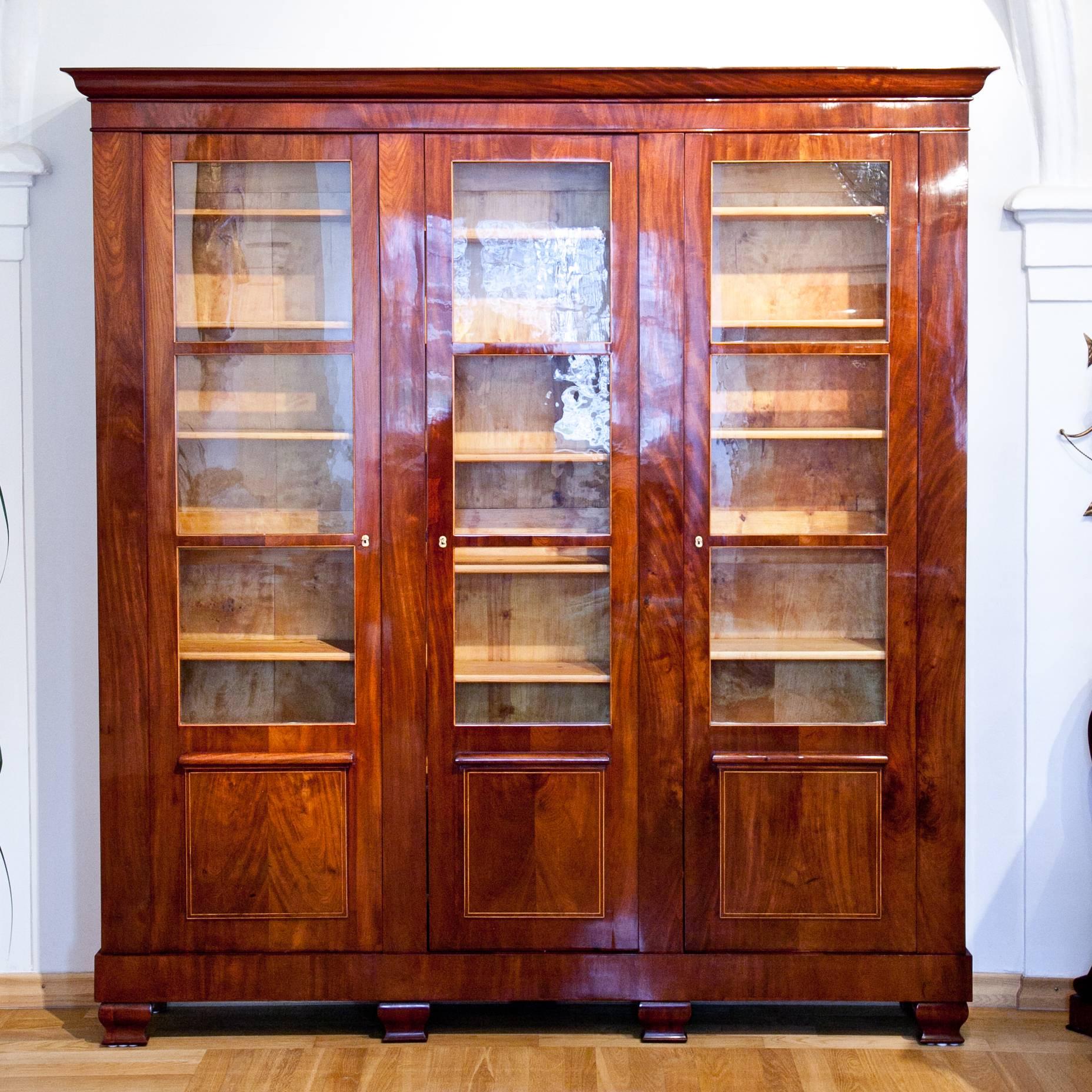Shallow library cabinet with three doors and old three-quarter glazing. The streamlined body is veneered with Mahogany, has a slightly accentuated base and a protruding cornice. The fillings are raised and framed, like the windows, with fine maple