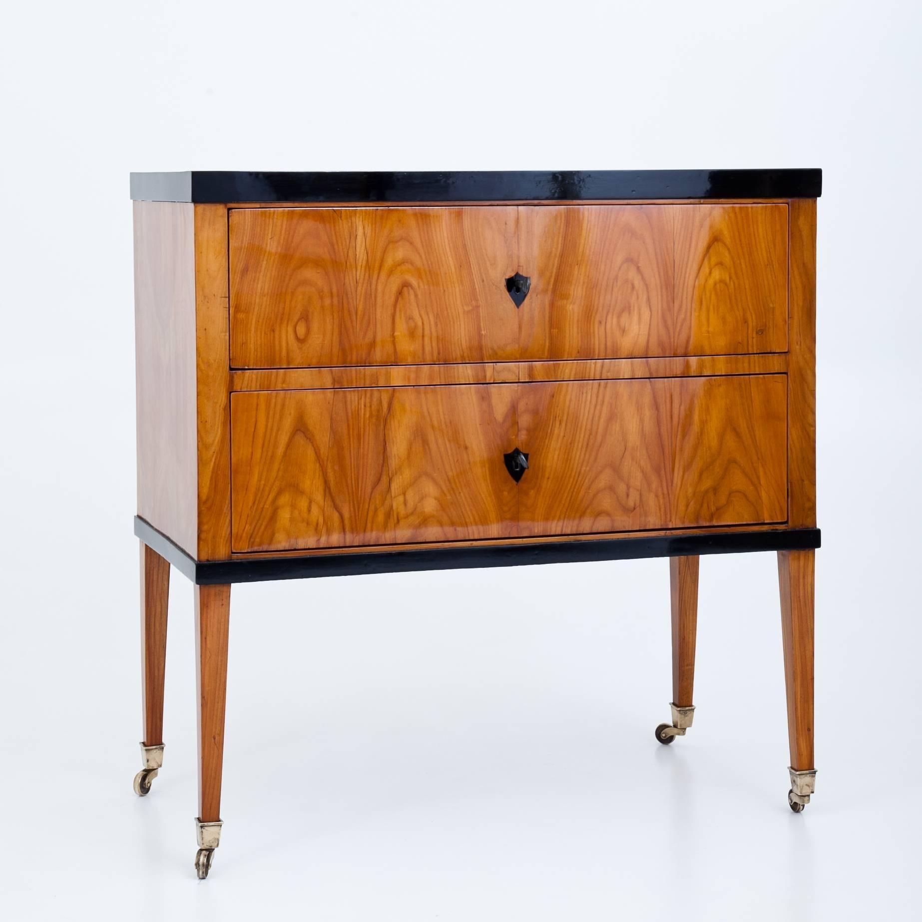 Two-drawered Biedermeier chest of drawers on long tapered legs with brass sabots on wheels. The edges and escutcheons are ebonized.
