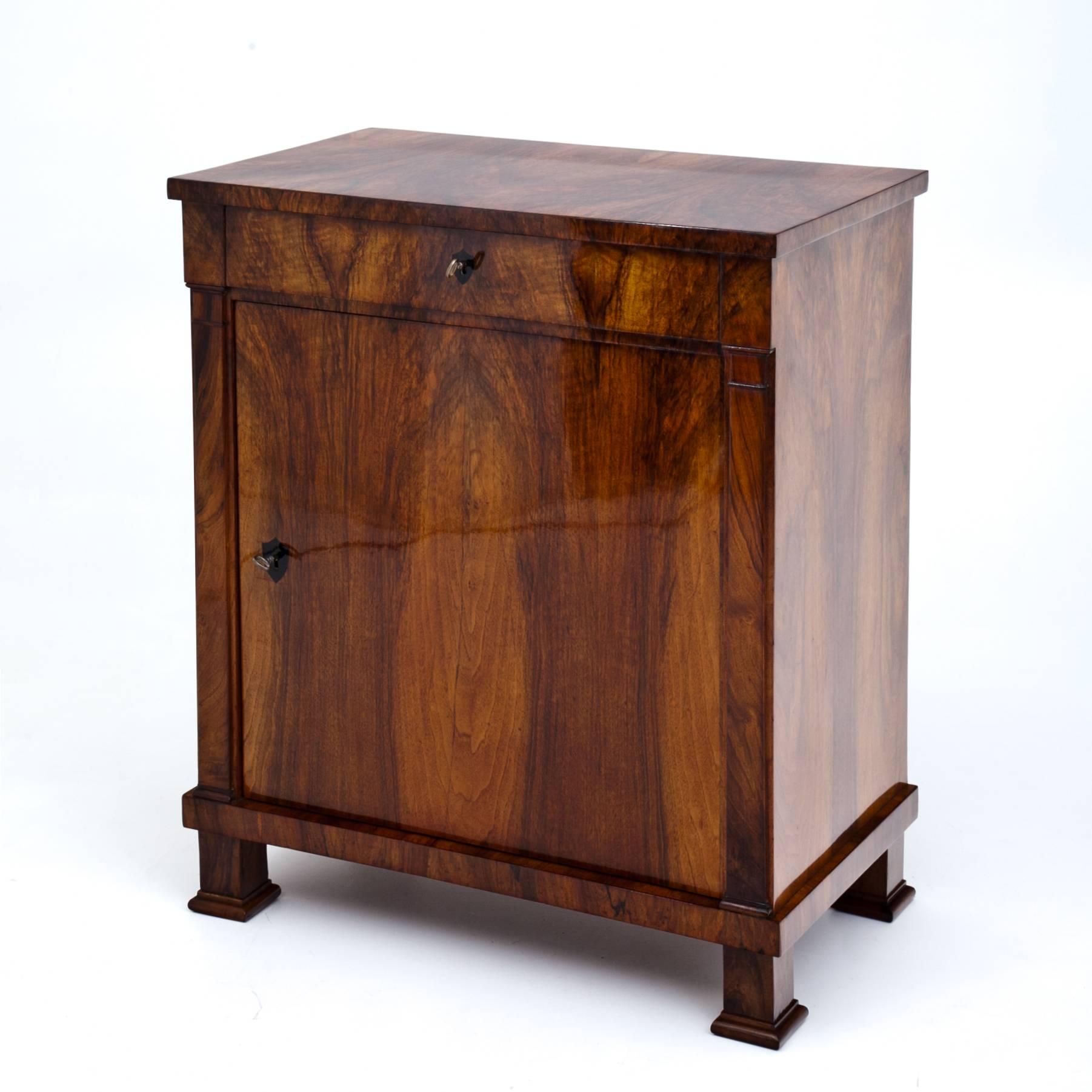 Small Biedermeier cabinet on square feet with small bases and one top drawer. The streamlined body shows a beautiful walnut veneer. A pair of pilasters frame the door and the locks are decorated with ebonized escutcheons.