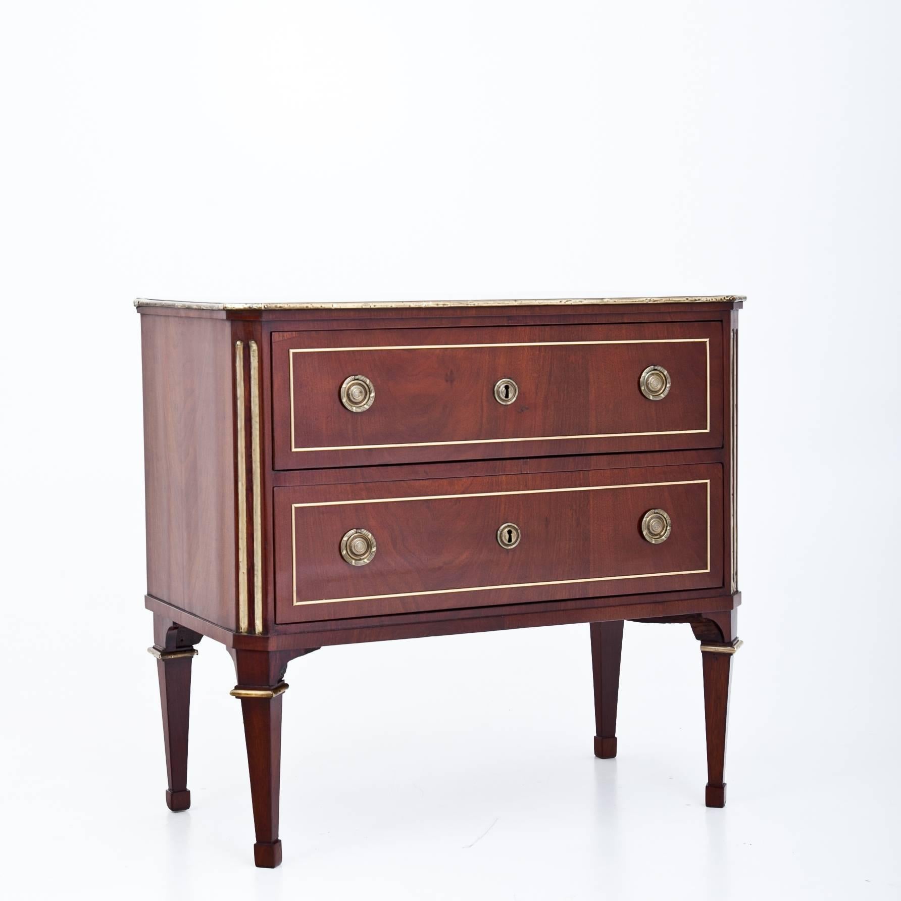 Two-drawered mahogany French chest of drawers on tapered feet with brass profiles. The corpus has slanted and fluted corneres and is decorated with brass strips.