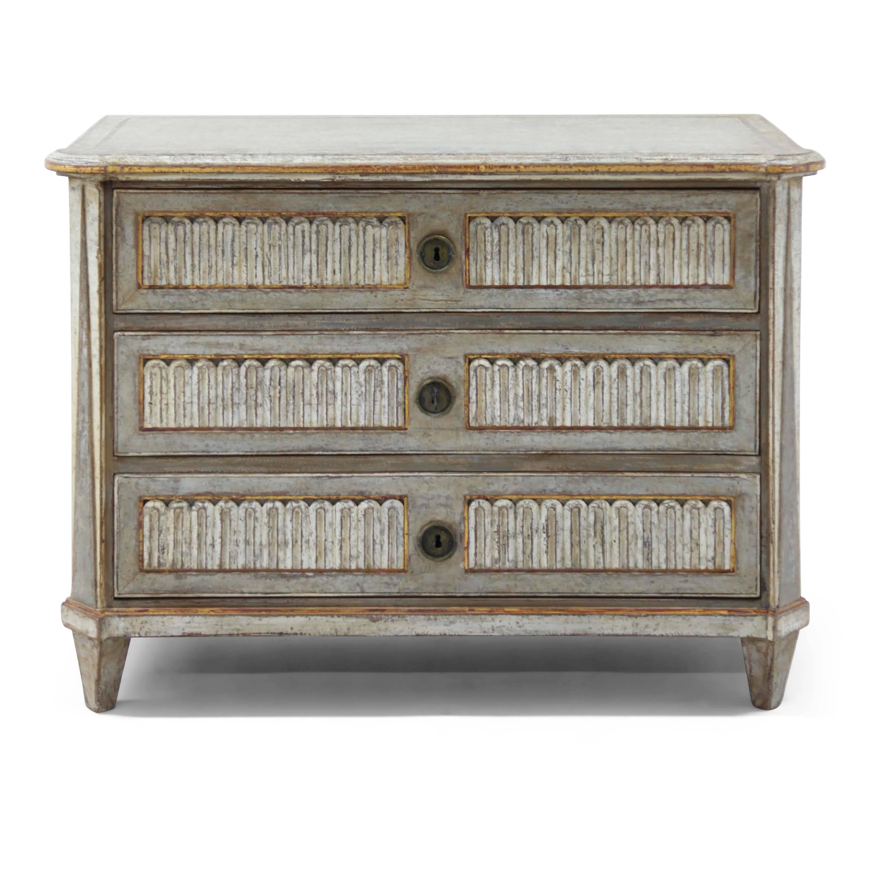 Three-drawered neoclassical chest of drawers with slanted corners, standing on tapered feet. The front and corners show carved details. The Gustavian style white and grey paint is new and has a worn look to it.