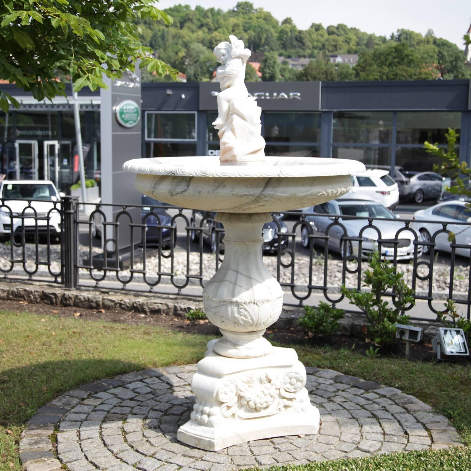 A 195 cm tall marble fountain with a cherub holding a fish in the middle. The fountain has many intricate details on the base and under the basin. The surface is very fine. This piece is a unique handcrafted fountain.