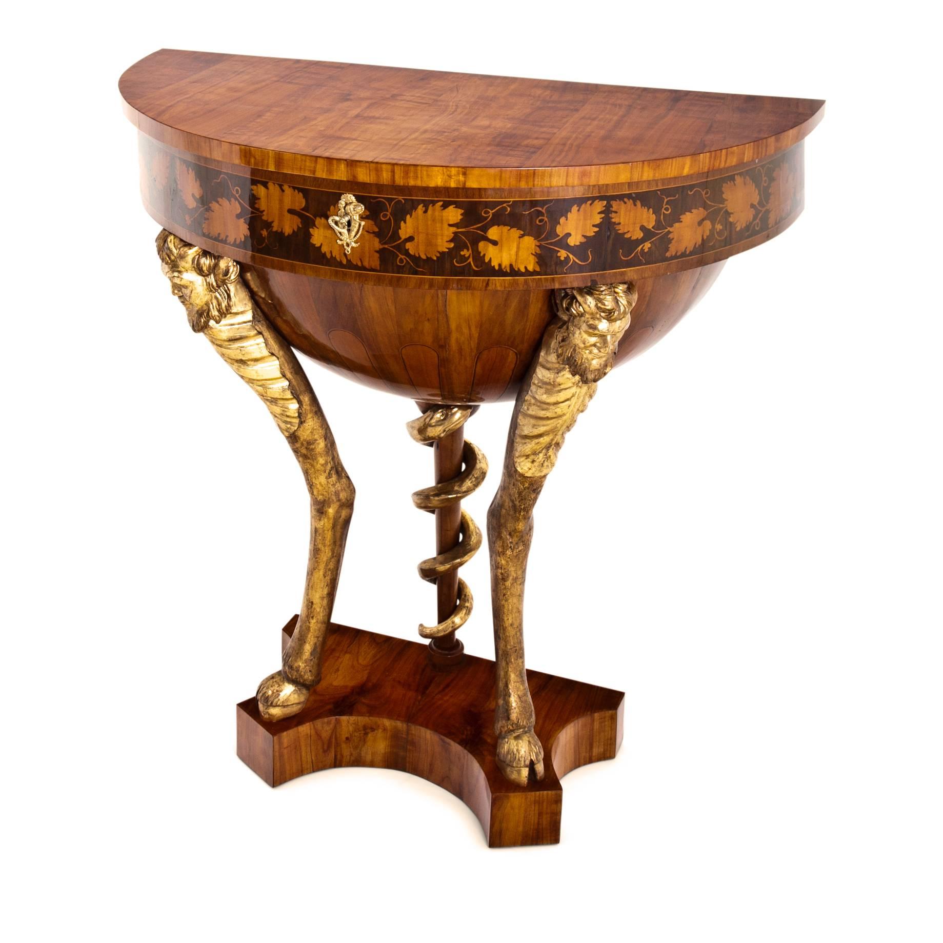 Empire console table on a trefoil base with legs in the shape of gilt satyrs with hove feet and one shaped like the rod of Asclepius with a gilt snake. The demilune body shows inlays in the shape vines at the front and holds one large compartment.
