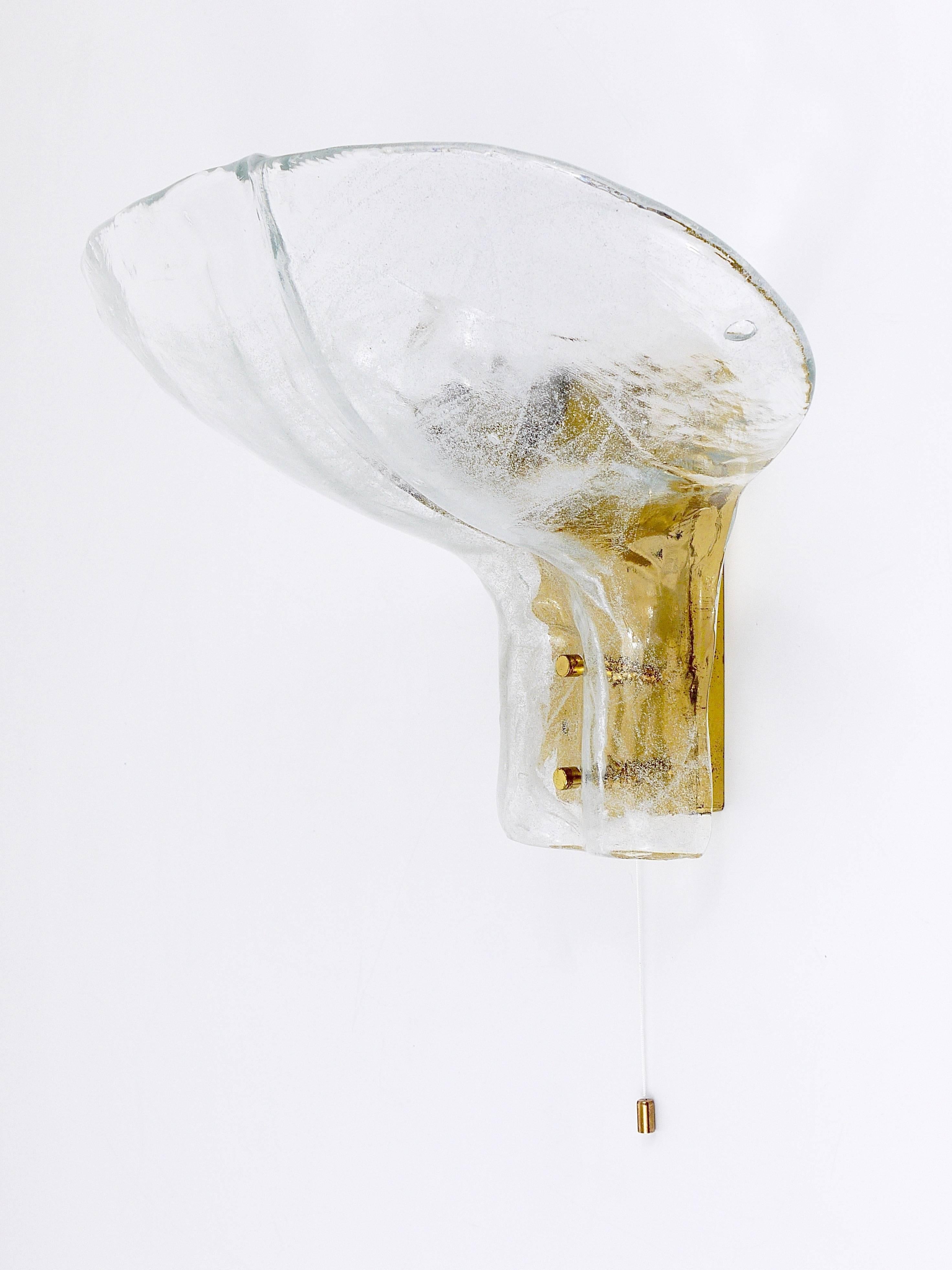 A sizable Mid-Century brass wall light with a blossom-shaped ice glass panel and integrated switch. This exquisite piece was designed and crafted by J.T. Kalmar in Vienna, Austria during the 1970s era. The lampshade is made of thick handblown clear