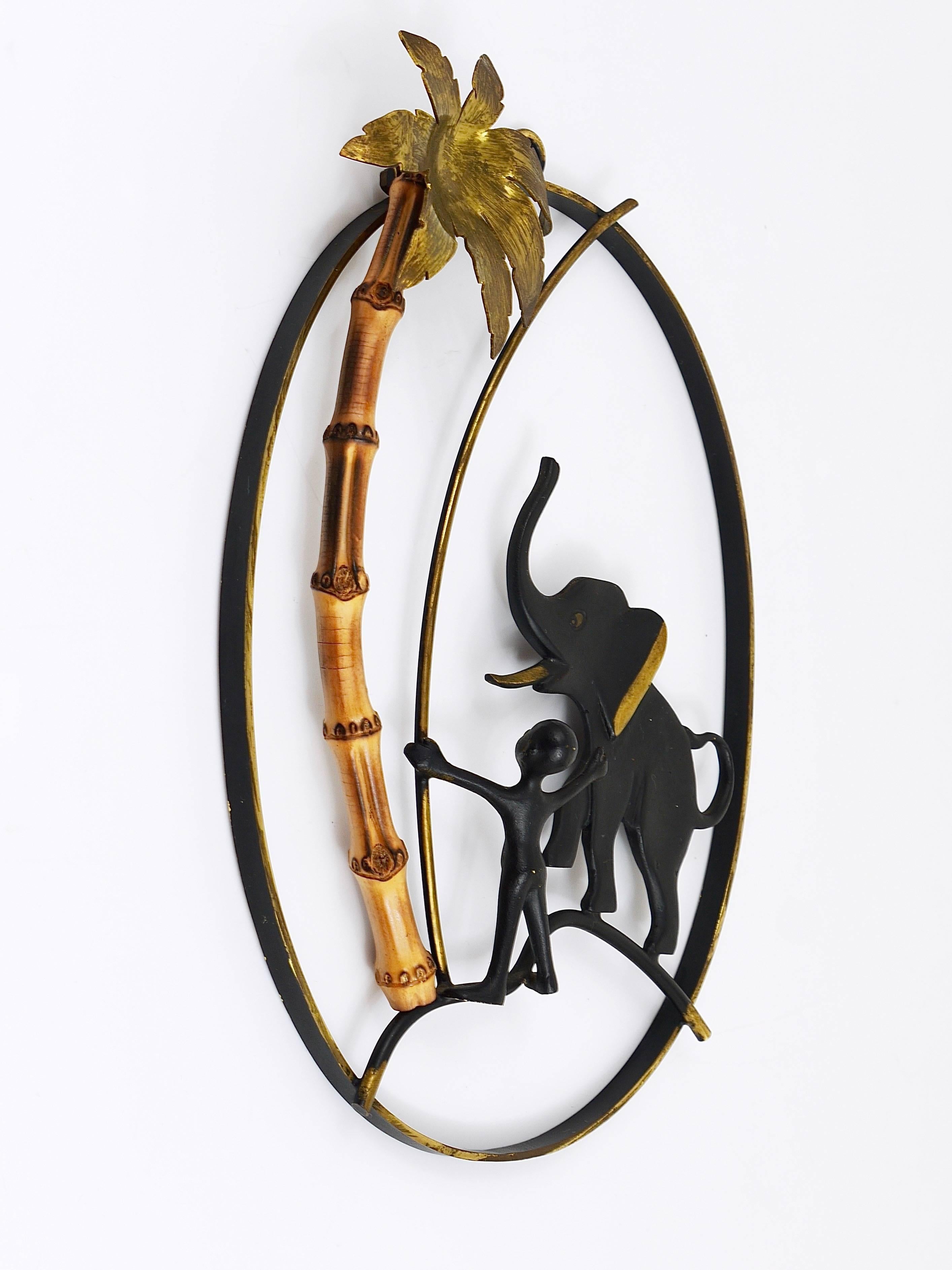 A lovely Mid-Century wall-mounted brass sculpture, displaying an African and an elephant under a palm tree on a frame, made of black-finished brass and bamboo. Designed by Walter Bosse, manufactured in the 1950s by Hertha Baller, Austria. In