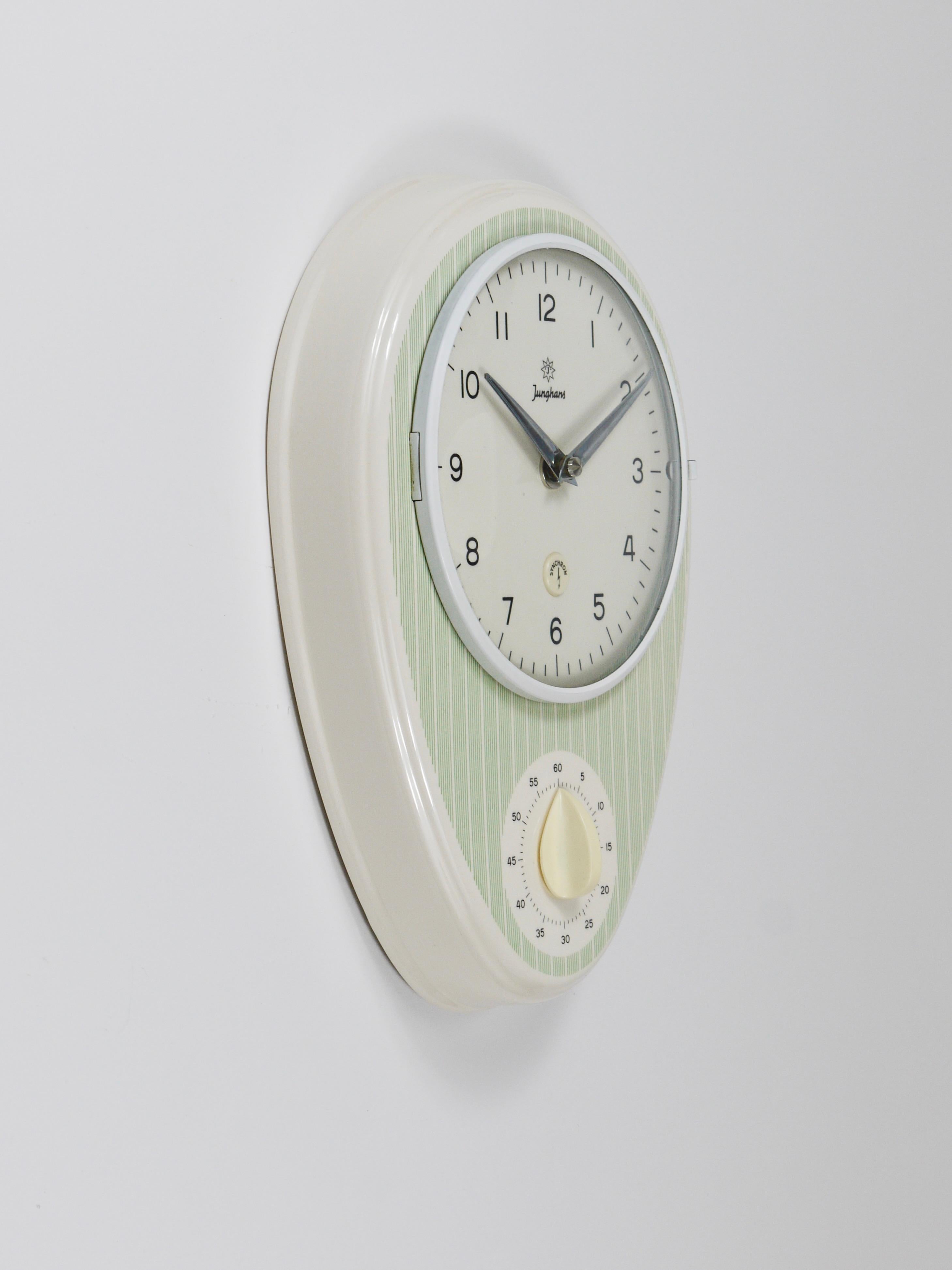 A lovely oval mid-century modern wall clock and timer, crafted by Junghans in Germany during the 1960s. Designed in the characteristic style of the era, it features elegant chrome-plated hands, a domed glass cover, and a white ceramic housing with