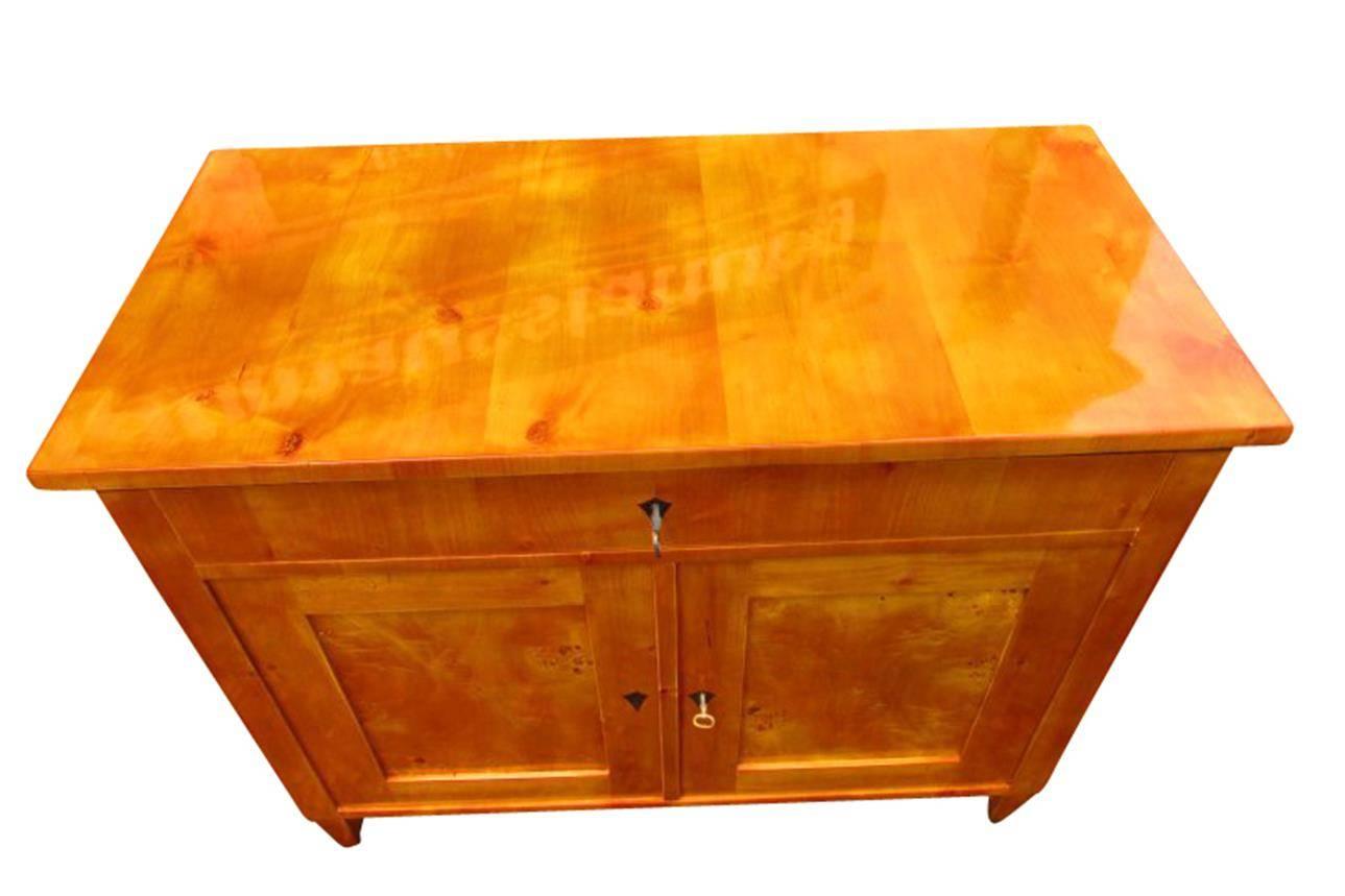 This rare commode from the Biedermeier period impresses with its honey colored birch veneer. The surface of the golden furniture is shellac polished.

Two doors, one drawer
Two keys and two locker
Classicly restored furniture with great