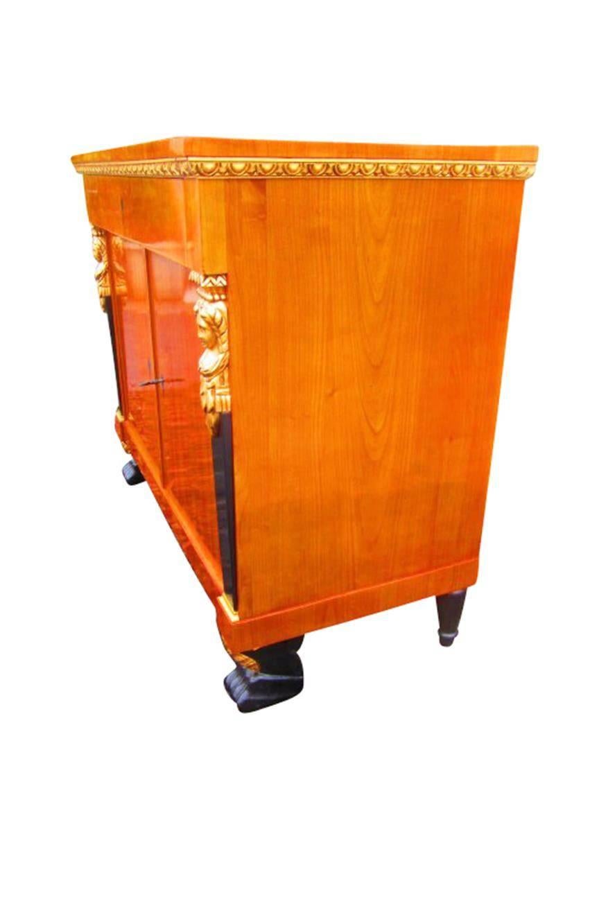 This aesthetic commode from the Empire period is made of massive cherry tree wood. The counter top and the doors are made of cherry tree wood veneer. Both sides are decorated with gracile, golden women figures.

Shellac polished
Paw-feets
Golden