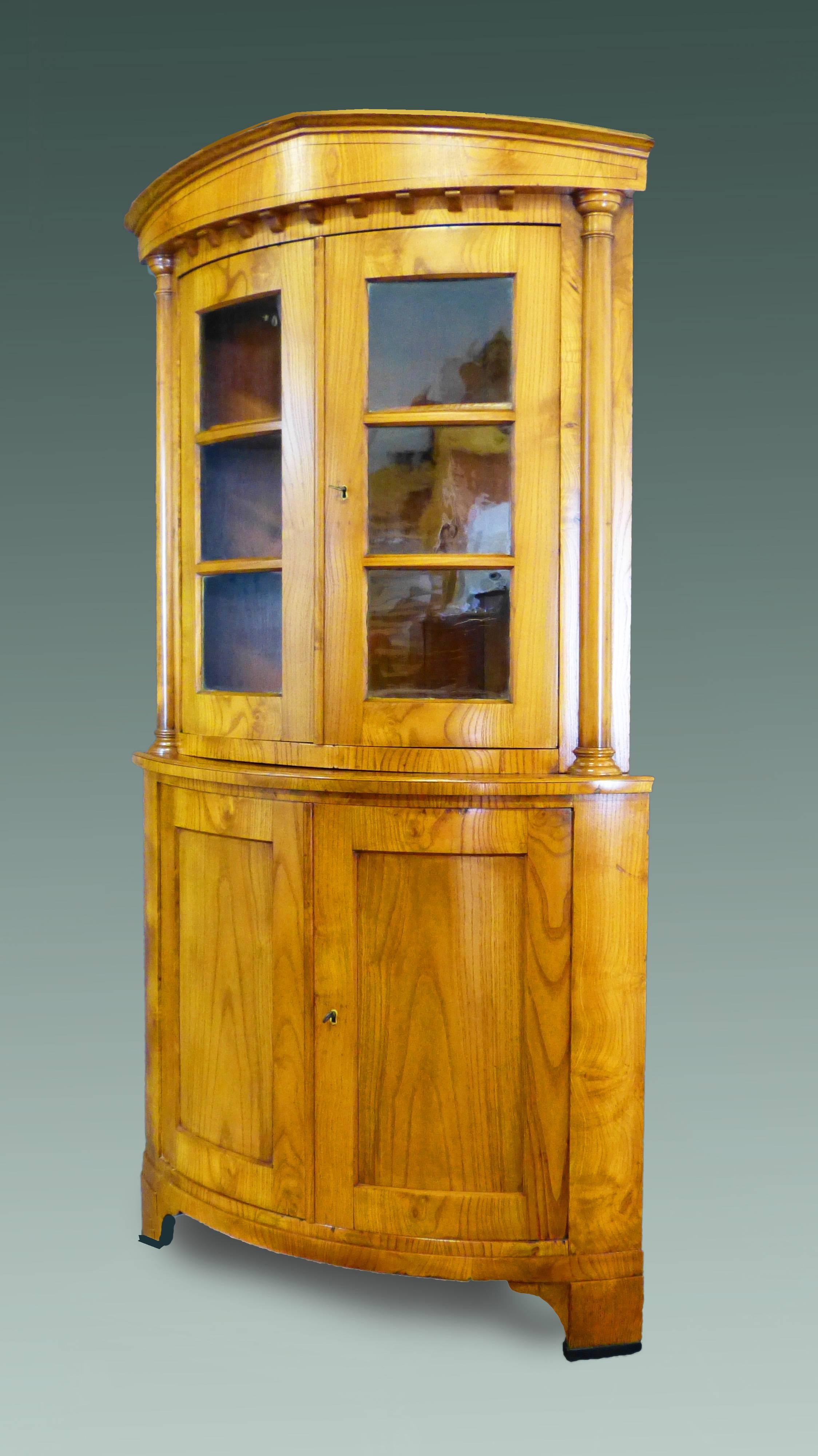 Rare early 19th century Biedermeier bow front corner cabinet of north German origin made of ashwood veneers over pine interior. The upper section with helmet top pediment with interrupted moulding below, two frontal columns flanking two curved doors