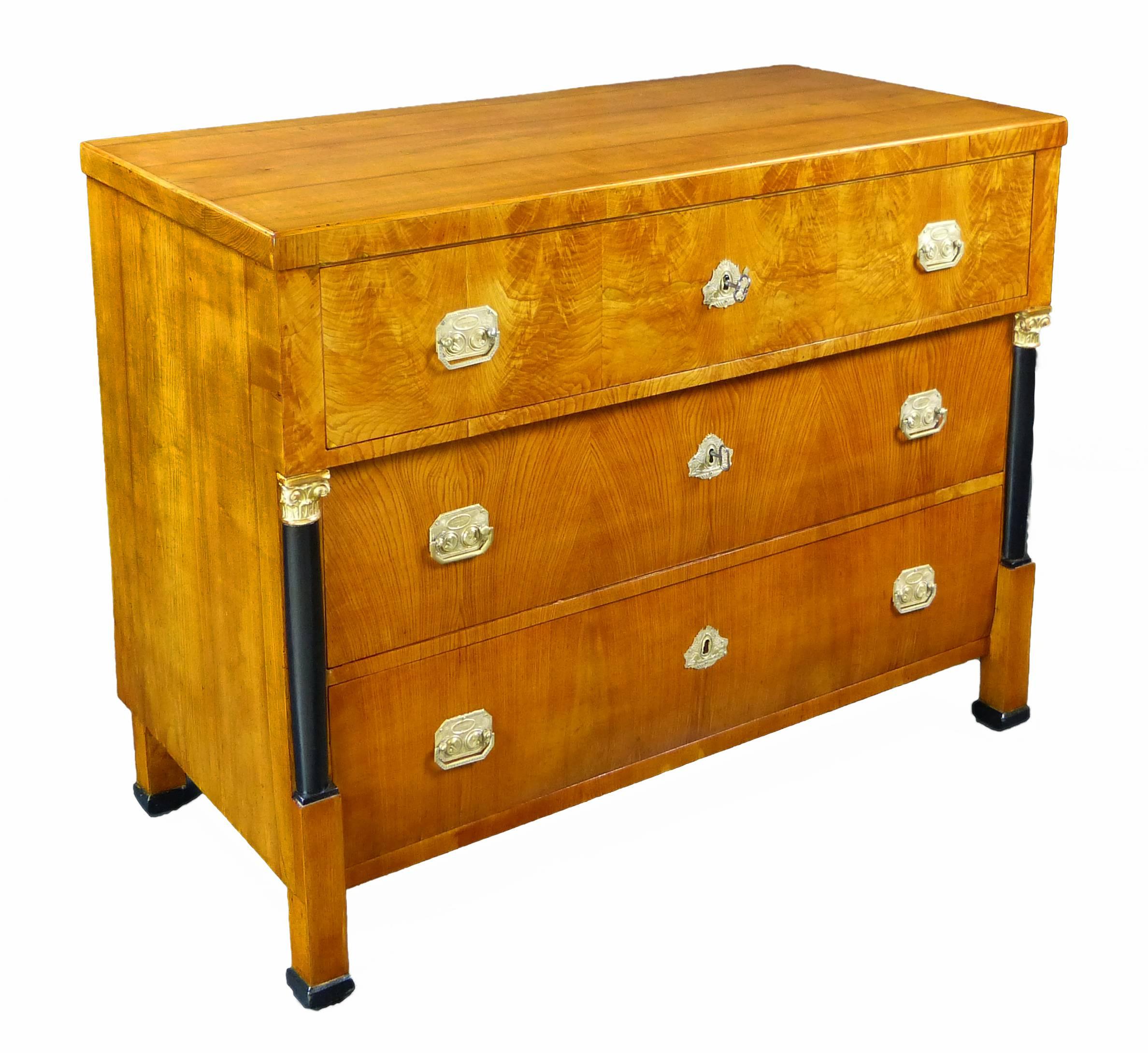 Fine late Empire early Biedermeier commode of German origin made of elm and burr elm veneer. There are 2 frontal ebonized columns with giltwood Ionic capitals. The 3 graduated drawers feature commemorative pressed brass pulls that feature 2