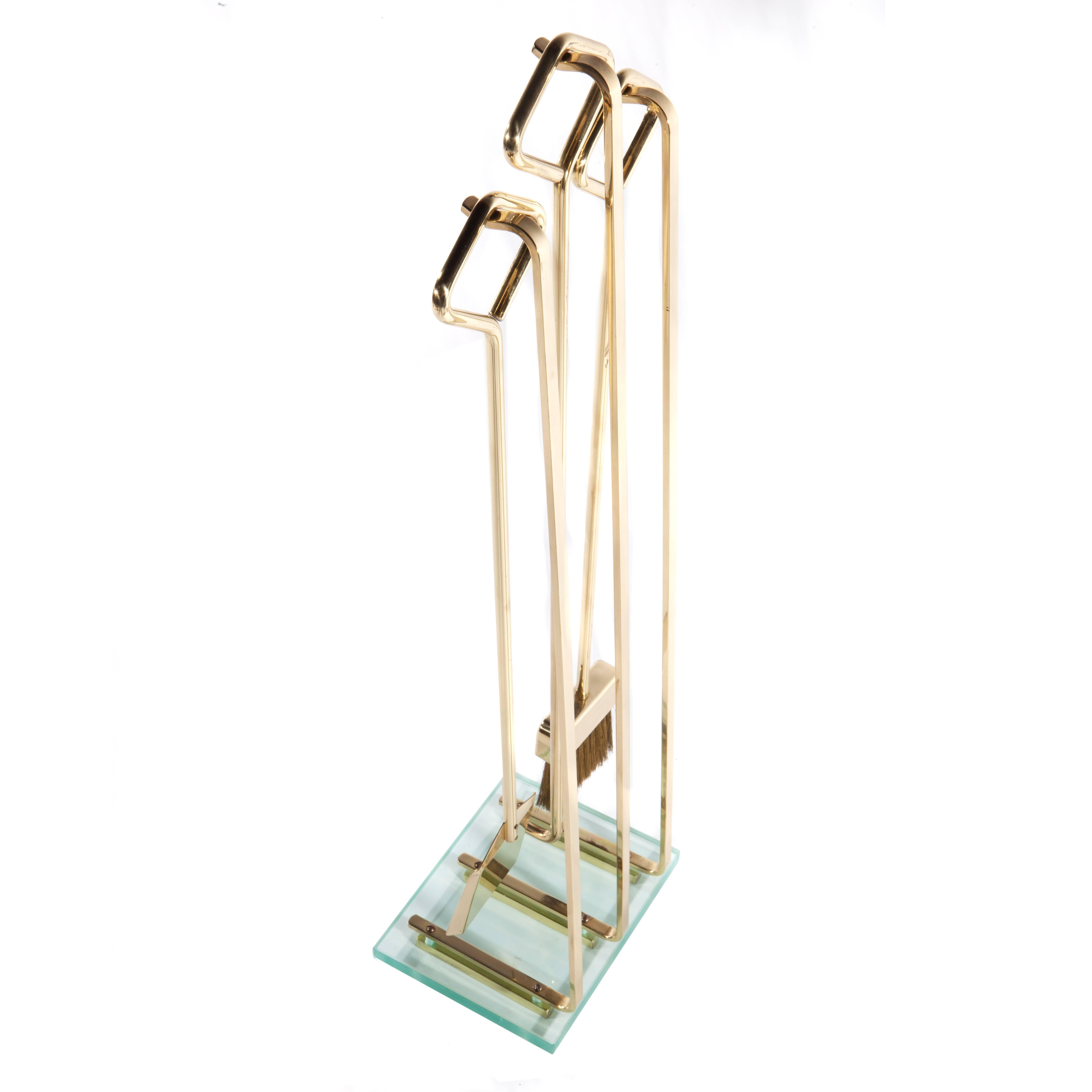 1970s fireplace tools with shovel, poker and broom, each ending in a diamond-shaped, bent-brass handle. Thick, rectangular glass base anchors three brass support hooks. 


