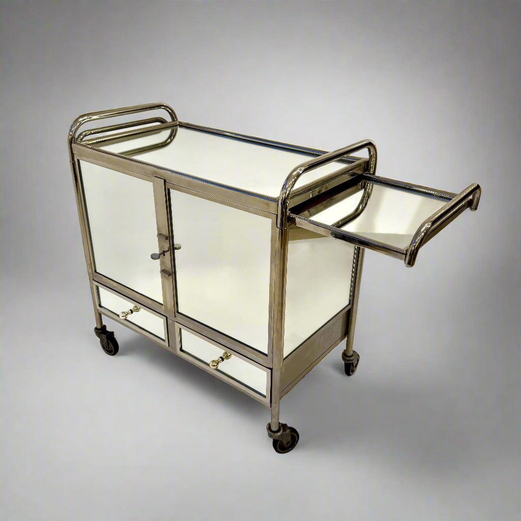 An Art Deco bar cart, made of polished nickel frame with mirrored surfaces and pull out extension trays on either side. The interior is fitted with bottle and glass holders. At the bottom are two drawers for storage as well.