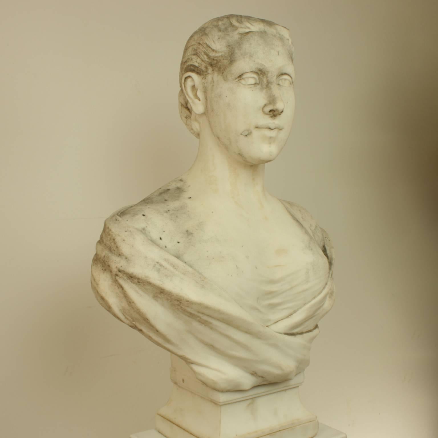 The marble bust facing frontal with her eyes looking to her right. The hair fastened in a skillfull knot behind, the drapery covers her shoulders and breasts, with another section underneath. The young woman displays an observing gaze and knowing