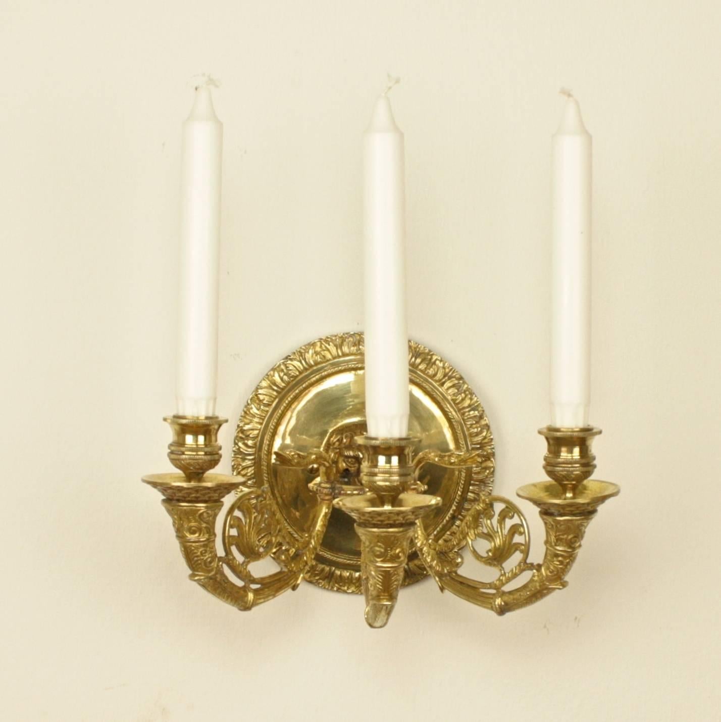 Pair of finely cast gilt-bronze wall lights, each with a circular back plate with a central lion's mask holding a ring issuing three swan neck outscrolling candle-arms with circular stiff-leaf decorated drip pans. The pair can be fitted for