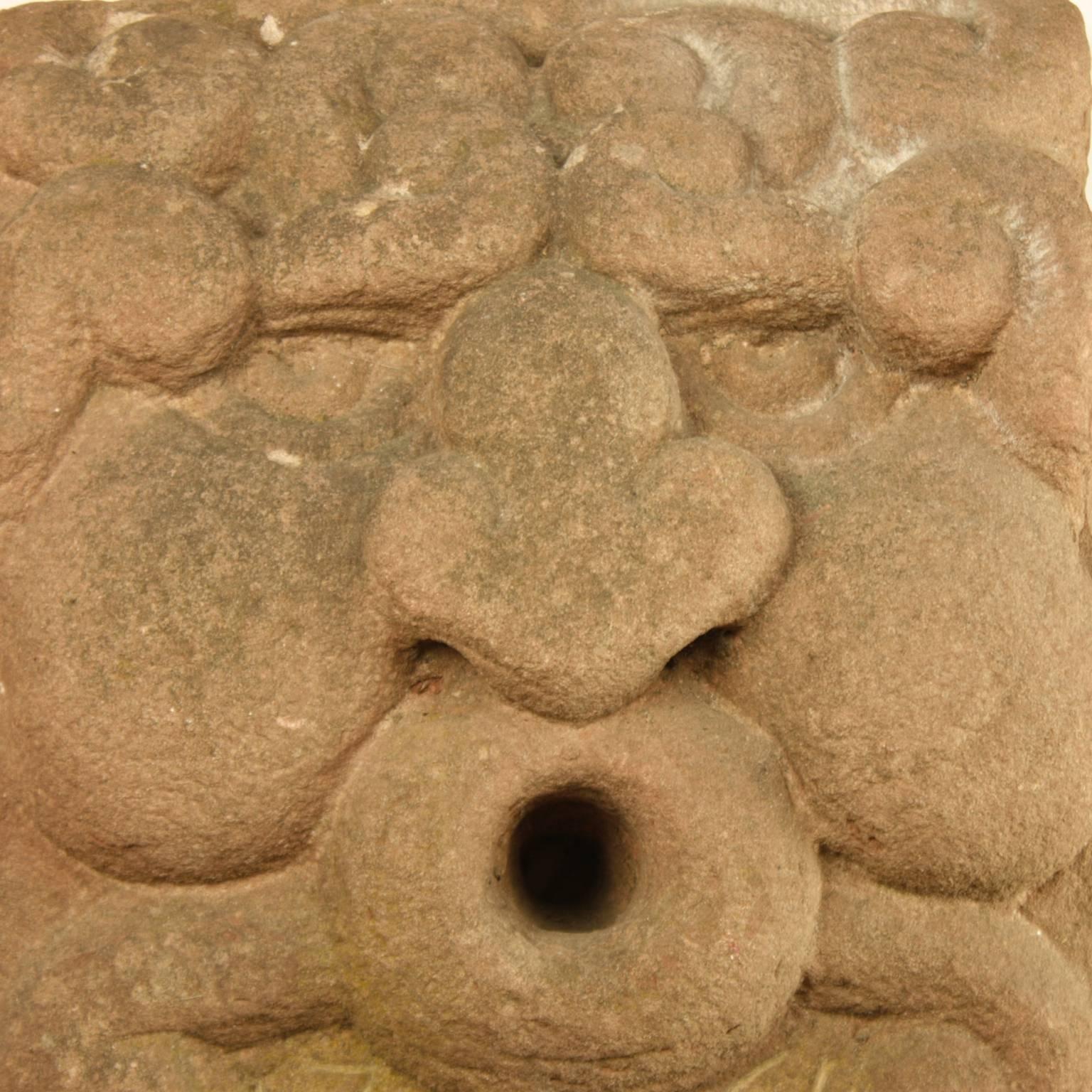 An 18th century red limestone gargoyle head, depicting a water deity, most likely of the river and functioning as water spout. The head with bulging nose, cheeks and open mouth inscribed 