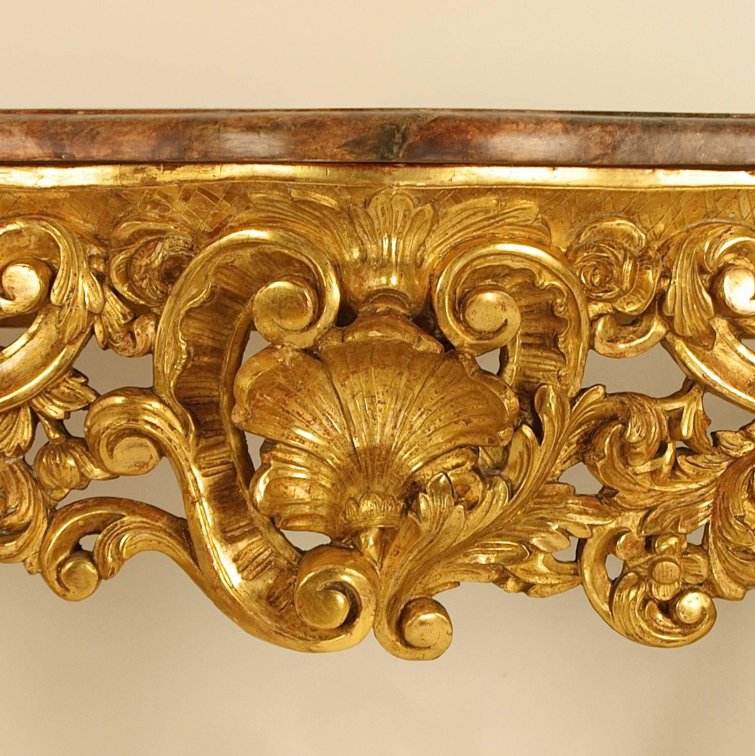 A giltwood carved console table with a faux mottled rouge marble top of serpentine form, above a pierced frieze boldly carved with a central shell within C-scrolls, acanthus leaves, rocaille and flowers. Raised on cabriole front-legs joined by a