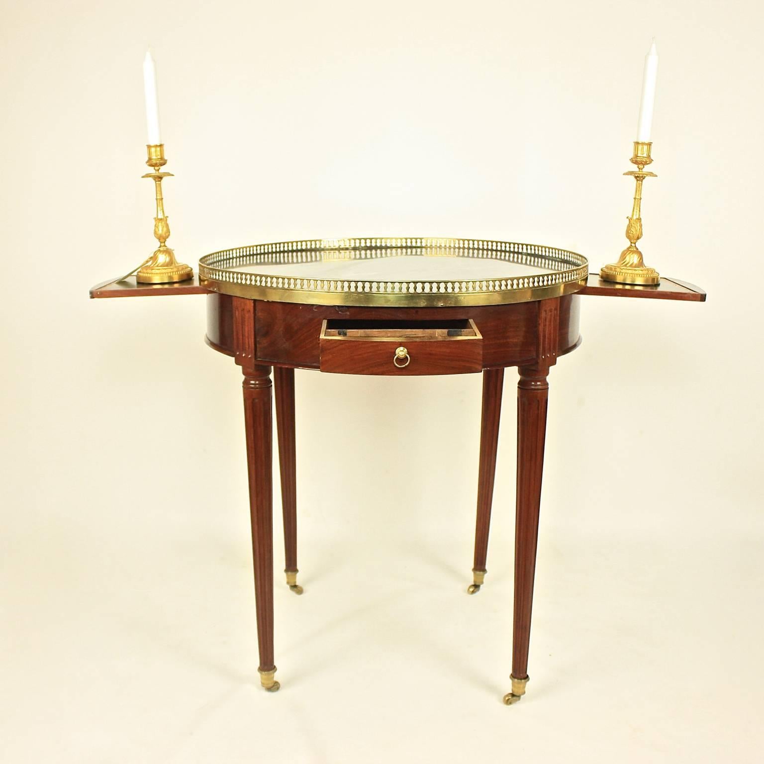A late 18th century mahogany bouillotte table, the circular grey-veined marble top with a pierced gallery above two frieze drawers flanked by two leather-lined candle slides, with the well-preserved removable top displaying a leather-lined playing