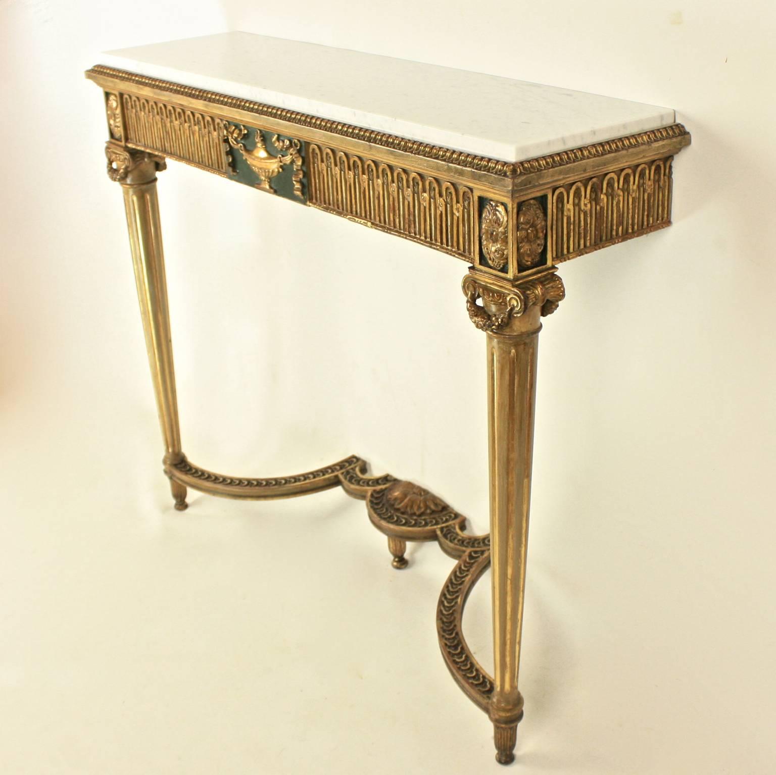 A pair of narrow Louis XVI style console tables, each with a rectangular white marble top above a fluted frieze centered by a ribbon tied giltwood urn on a dark green background and paterae on the corners. The tables are raised on fluted tapering