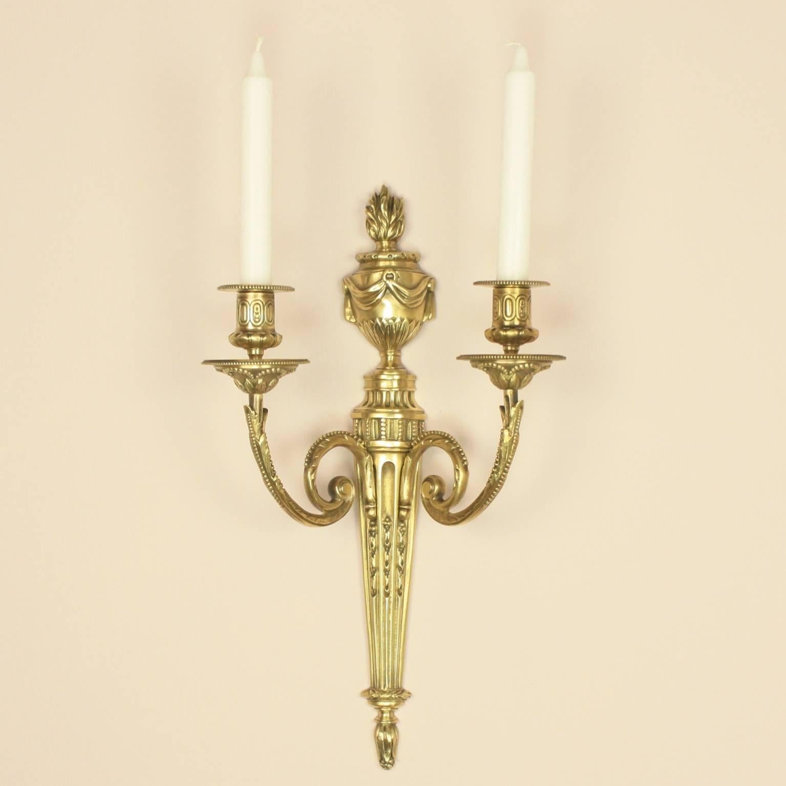 A pair of 19th century bronze wall-lights, each with a fluted back plate surmounted by a swagged flaming classical urn, with two scrolled candle arms cast with leaves and beading and foliate cast drip-pans with corresponding nozzles, the fluted
