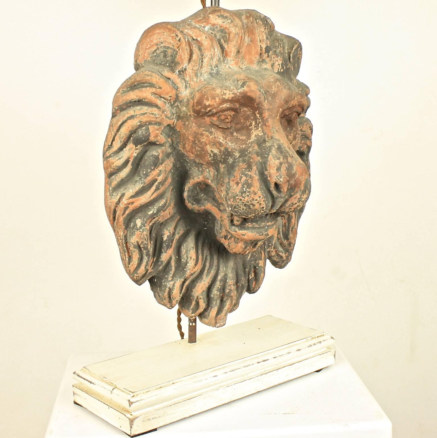 A pair of terracotta table lamps, each featuring an 18th century terracotta lion's head with remnants of a dark paint layer. The lion's heads have recently been converted to modern table lamps with a cream colored wooden base and oval cream-colored