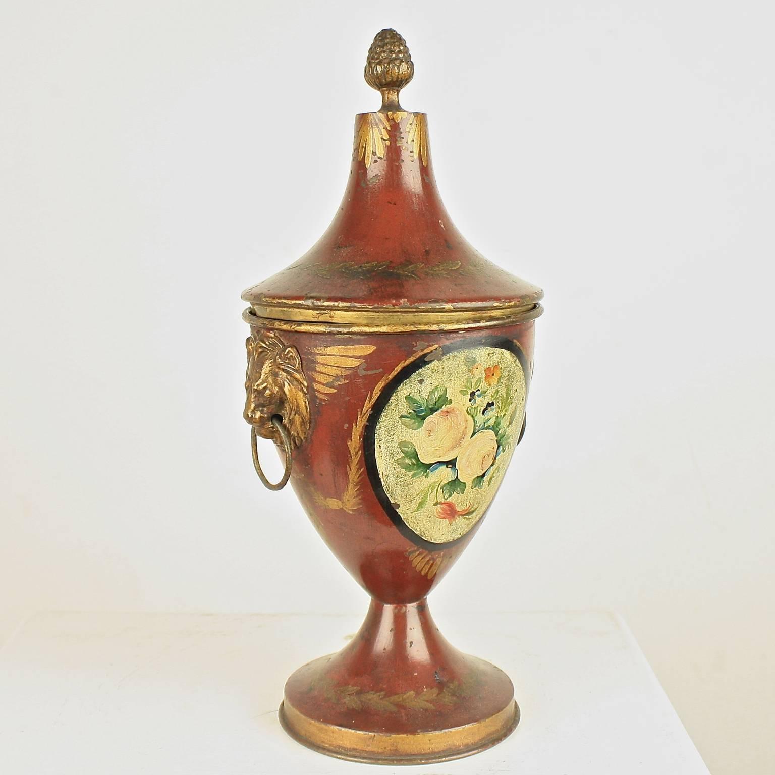 Early 19th century English Regency tole chestnut urn with a hand-painted floral decorative motif, a removable lid with acorn finials, original lions head side handles with rings, terminating on a circular molded edge plinths. The Venetian red body