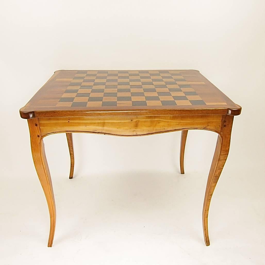 A French game table with a square top, canted corners and a removable round cornered game top displaying a chess game on top and a leather lined surface underneath. When the game top is removed the tric trac game is revealed, finely veneered in