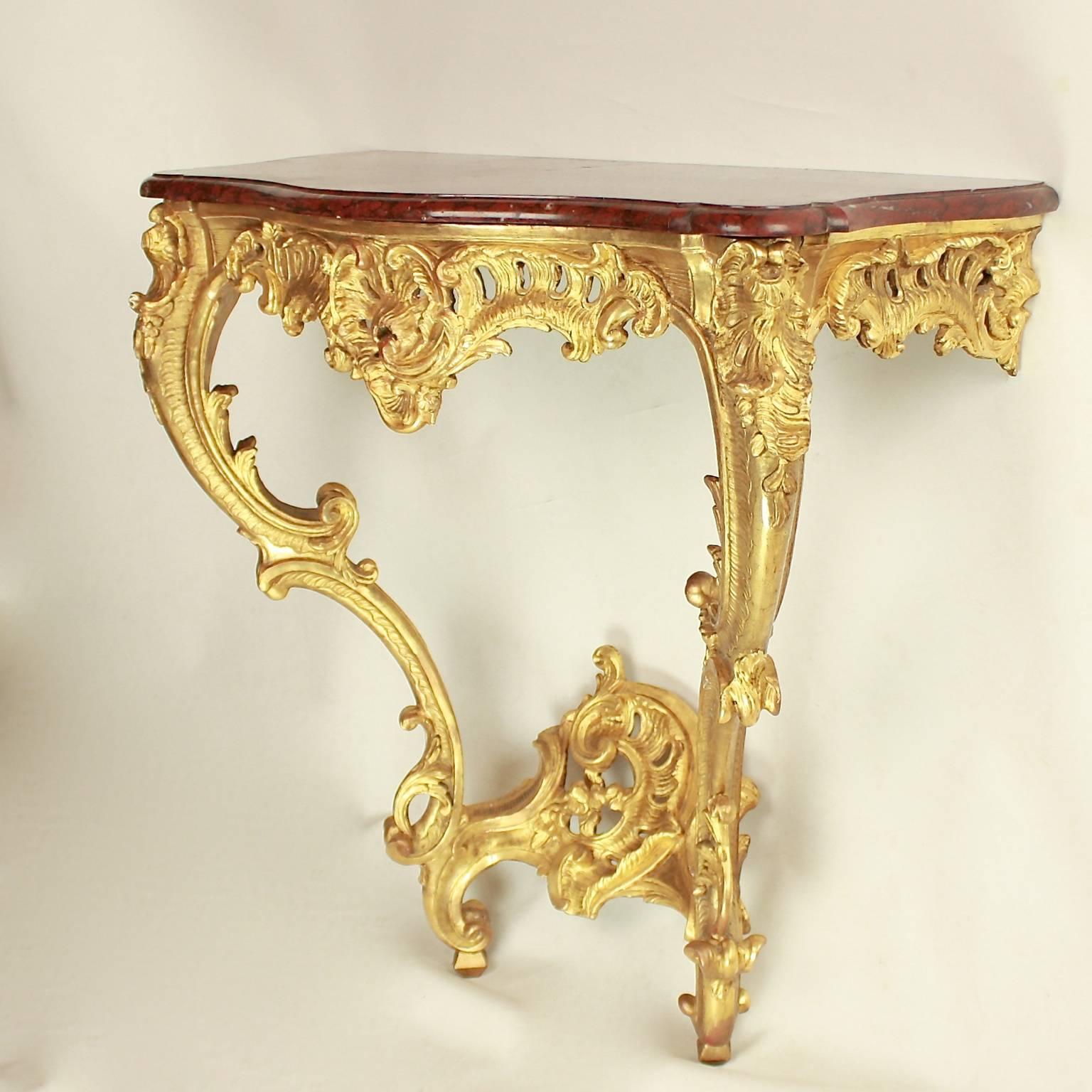 French Louis XV console table of finely carved and gilded wood. The red and grey 'rouge griotte' marble top of serpentine outline, with a pierced frieze carved with rocaille and leaves, with a stylised scallop shell on each corner, on cabriole legs