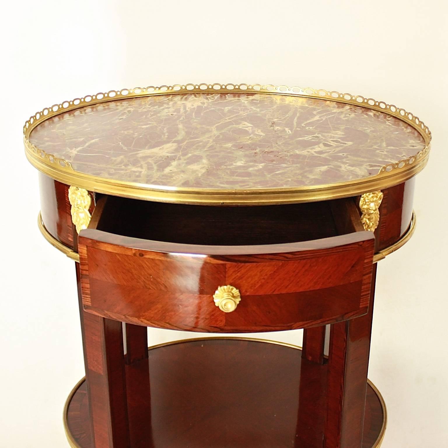 A Louis XVI style oval side table attributed to L’Escalier De crystal. The two-tier table with a dark red and white veined marble top and a gilt bronze gallery all around, the lower tier with brass banding, raised on cabriole legs. The frieze with