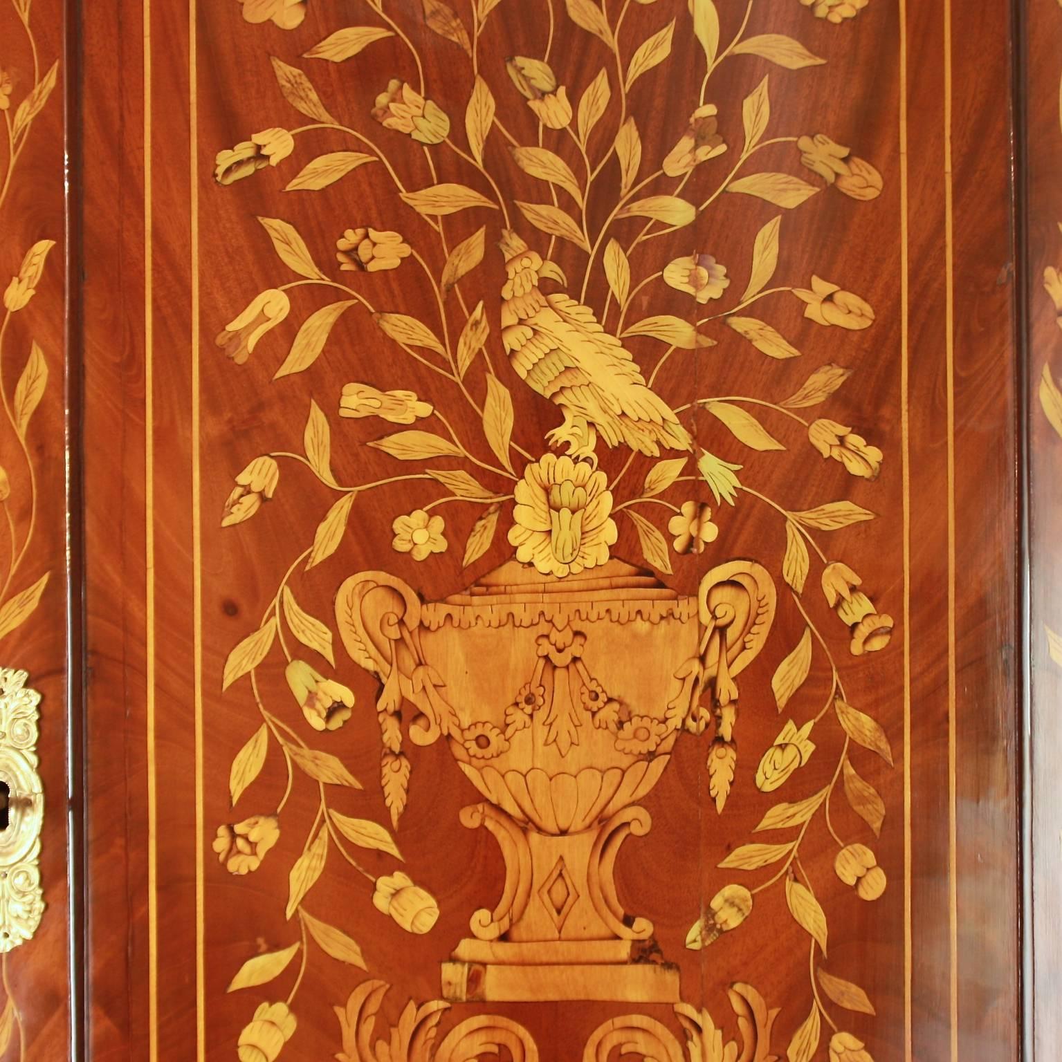 A large neoclassical bowfront corner cabinet of two parts in mahogany with floral marquetry in various woods (some stained), surmounted by a vase pediment with a classical urn. The top section with two doors displaying on each door a neoclassical