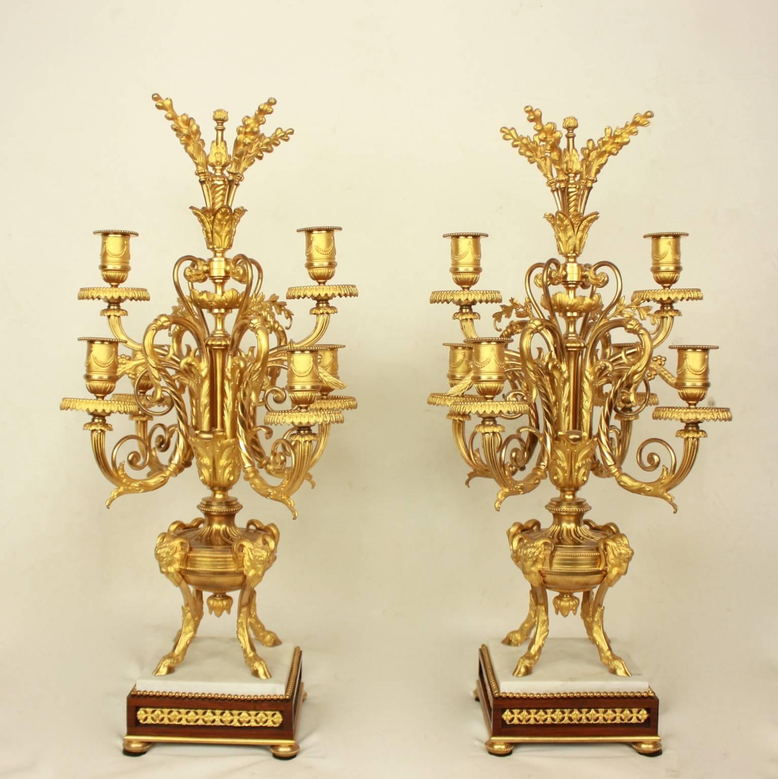 Pair of Louis XVI style ormolu candelabras, each with six scrolling candle arms and corresponding sockets and drip plates raised from a foliate cast central baluster. The central shaft emanating from a fluted urn supported by four rams on a square