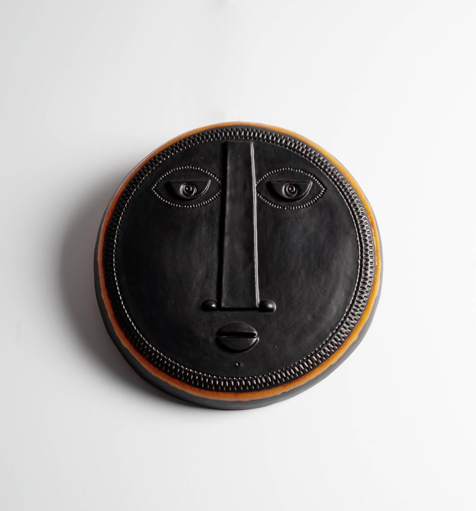 Decorative black ceramic mask with stylized face and shiny orange enamel around, creation 2017, unique piece by French ceramists Dalo.