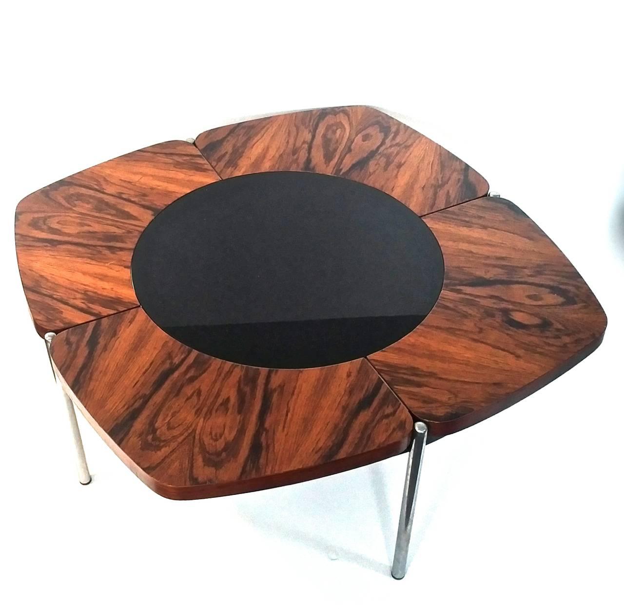 A Mid-Century coffee table with four sections forming a flower shape with a black glass center. Rosewood veneer on solid teak and a metal ring for support. The table stands on four chromed steel legs.