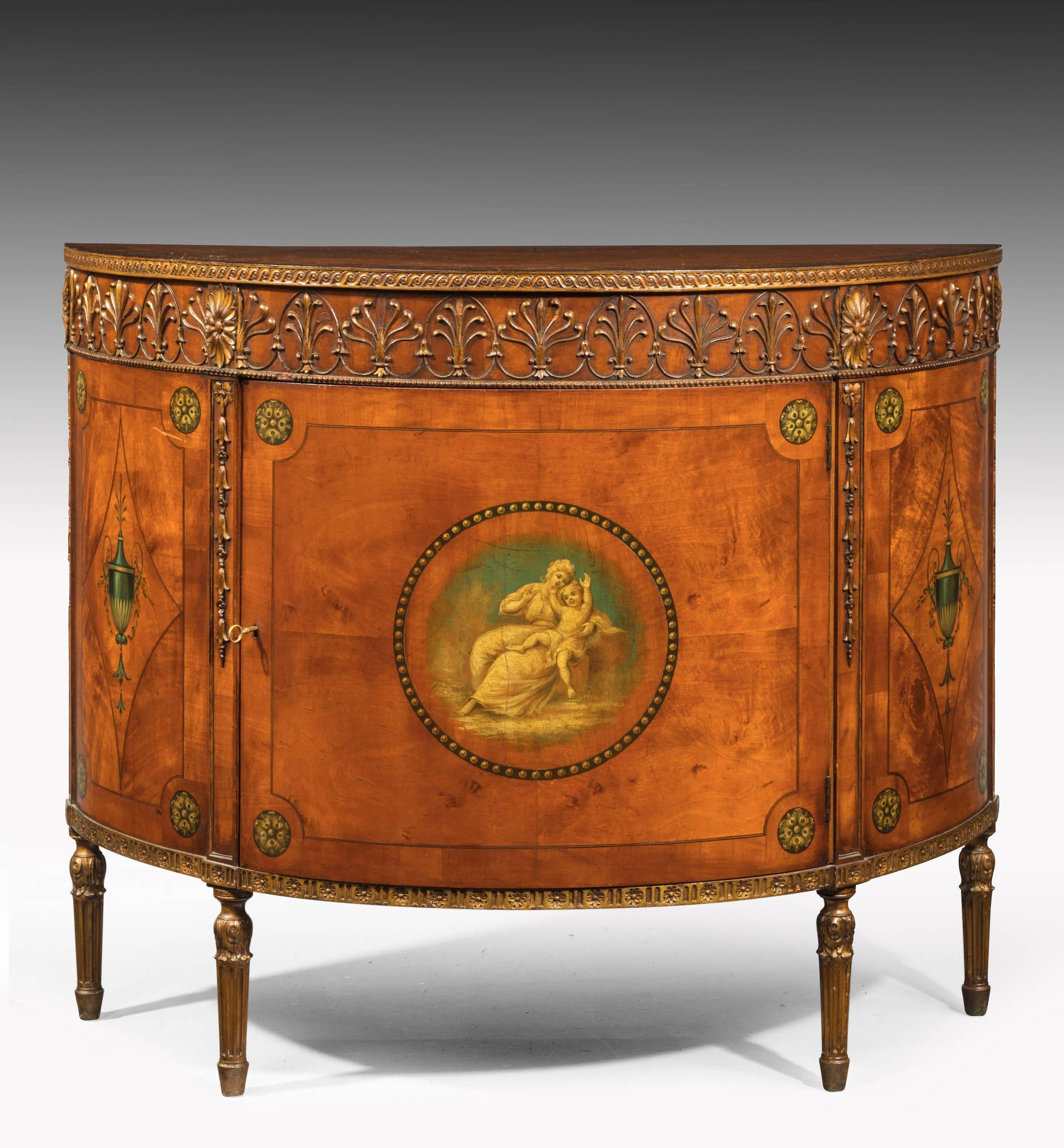 A fine pair of Sheraton style satinwood demilune Commodes. The top border beautifully carved with anthemion’s. The top edging painted and decorated retaining the original colors. The central motif of a neoclassical maiden with a child.