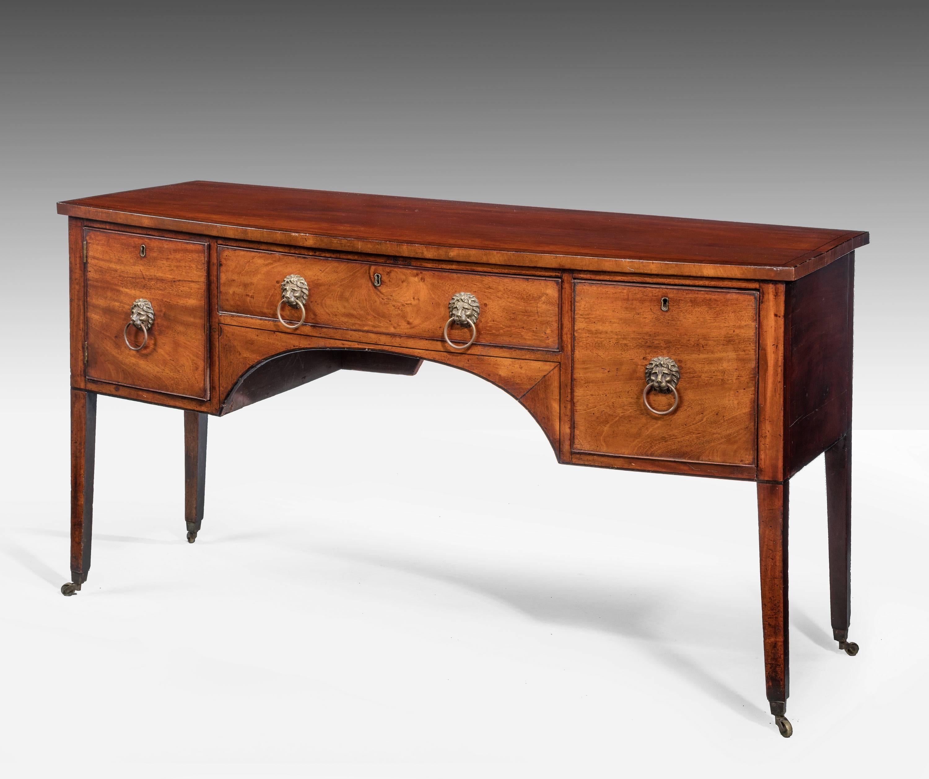 A George III period mahogany bow fronted sideboard, incorporating three drawers and on tapering square supports terminating in brass shoes and casters. Replaced but period lions mask handles beautifully cast. The top edge cross banded in matching