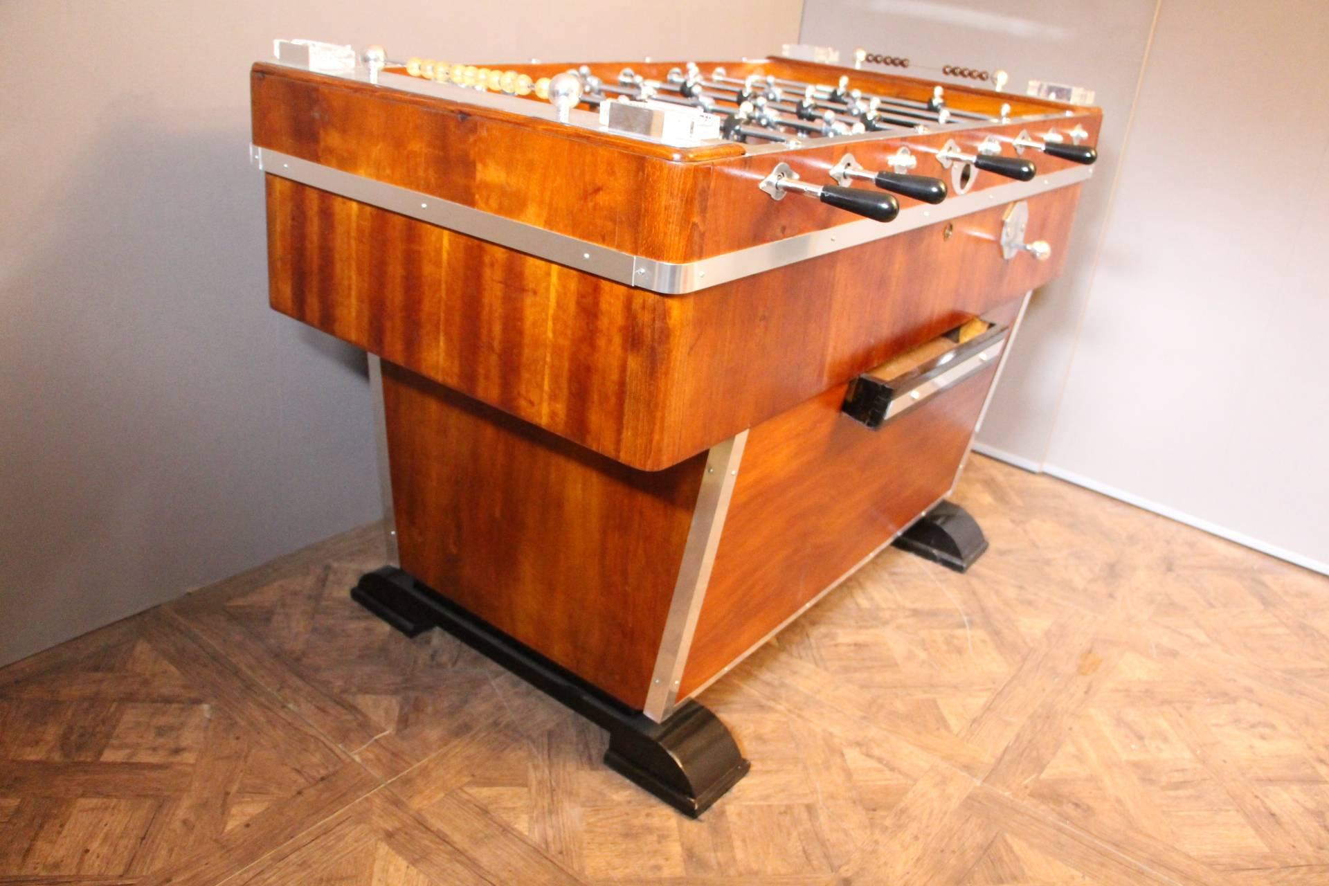 This foosball table is very unusual because it features a honey color walnut. It still has got its four original crystal ashtrays, as well as all its polished aluminium pieces. Its players are silver and black and the field is black. Its shape is